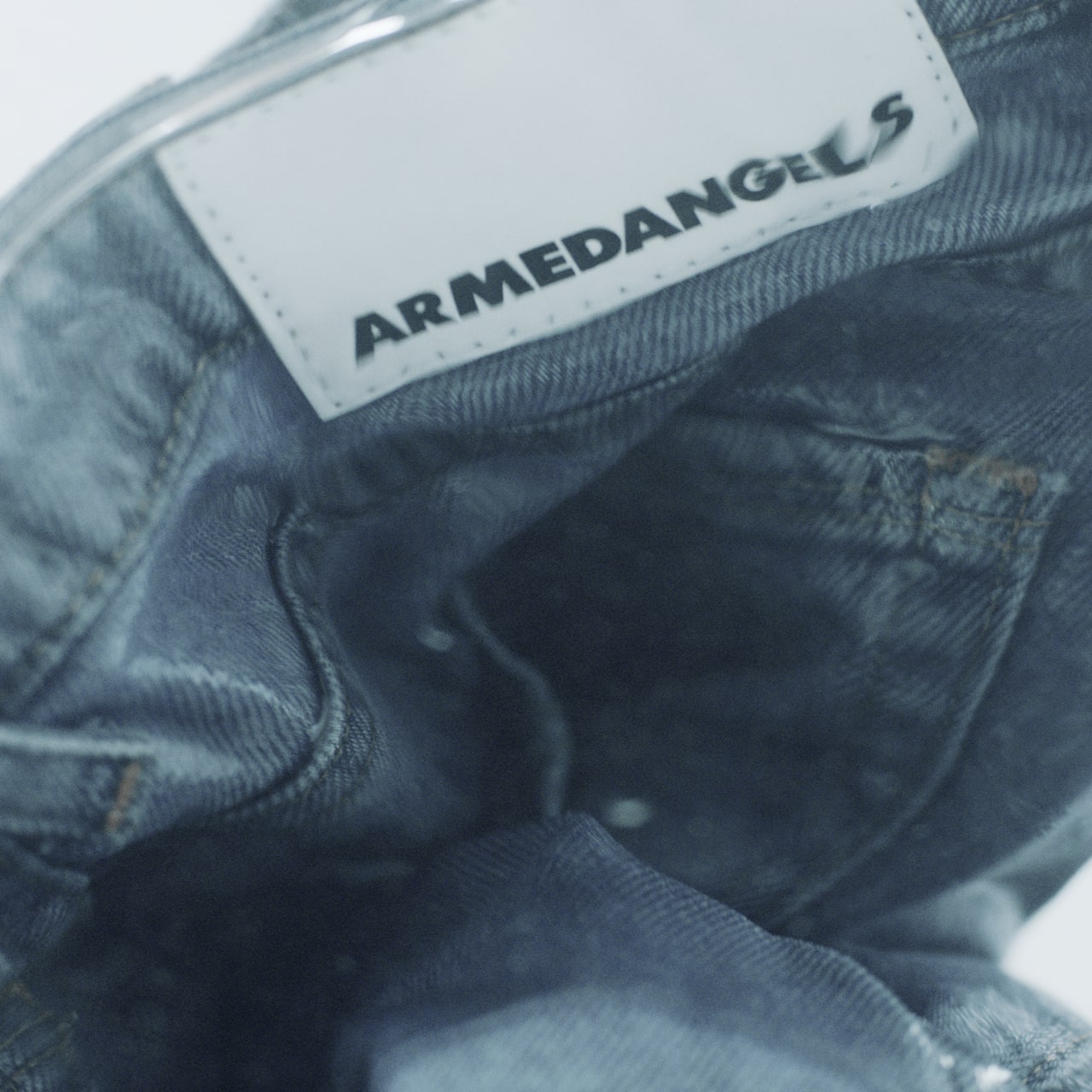 Nahaufnahme eines Jeansstoffs mit weißem Label, auf dem ‚ARMEDANGELS‘ steht. Das Etikett ist leicht geknickt, die Denim-Struktur im Hintergrund wirkt weich und leicht verwaschen.