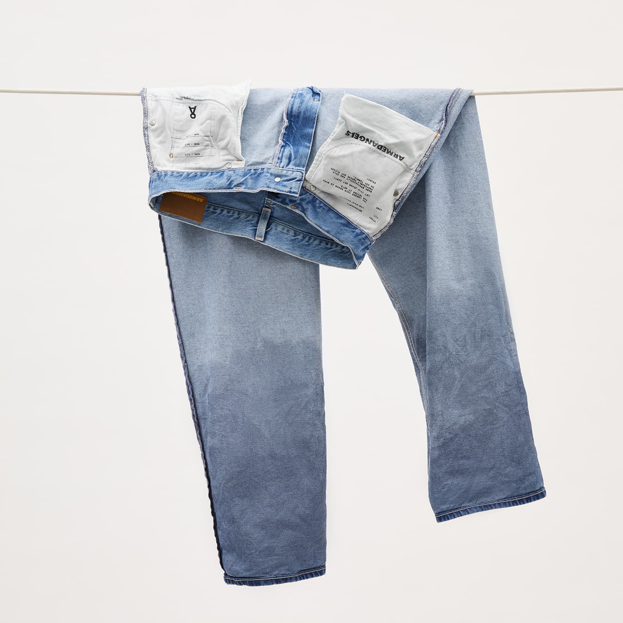 Eine aufgehangene Jeans in hell- bis mittelblauer Waschung hängt über einer Leine, auf links gedreht, sodass das Innenfutter mit ARMEDANGELS-Label sichtbar ist.