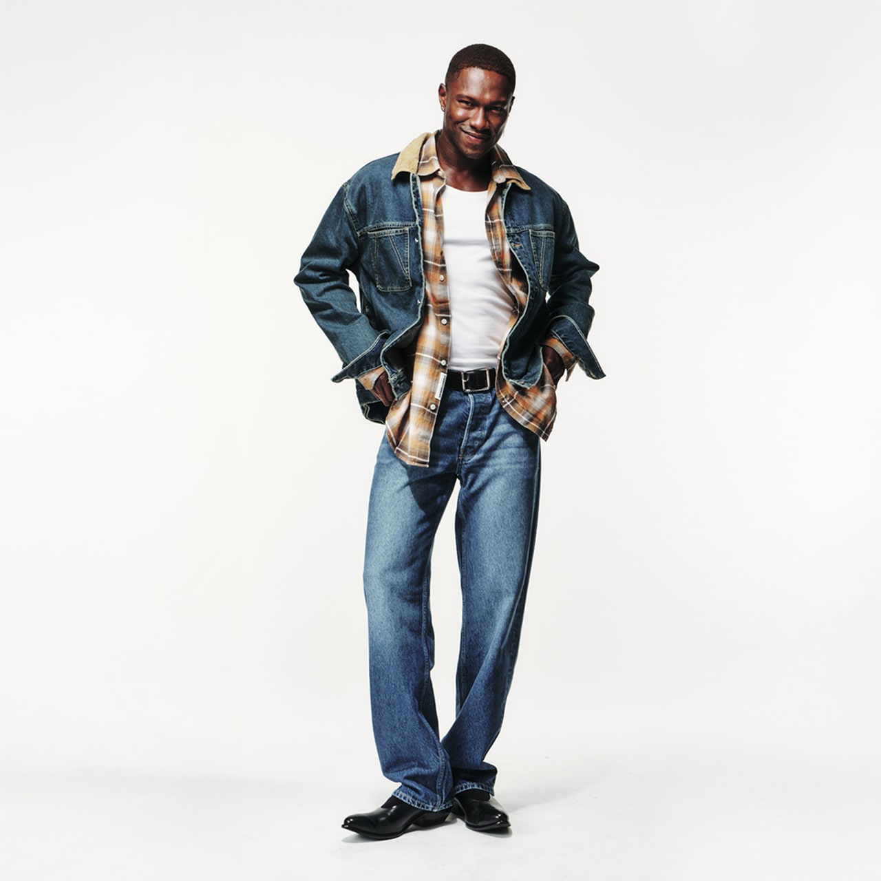 Person trägt Jeans-Outfit mit Denimjacke über kariertem Hemd und weißem Tanktop. Steht locker mit Händen in den Hosentaschen und lächelt leicht.