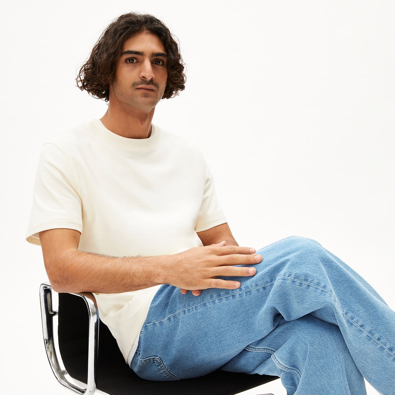 Person mit mittellangen dunklen Locken sitzt entspannt auf einem Stuhl, trägt ein cremefarbenes T-Shirt und eine hellblaue Jeans.