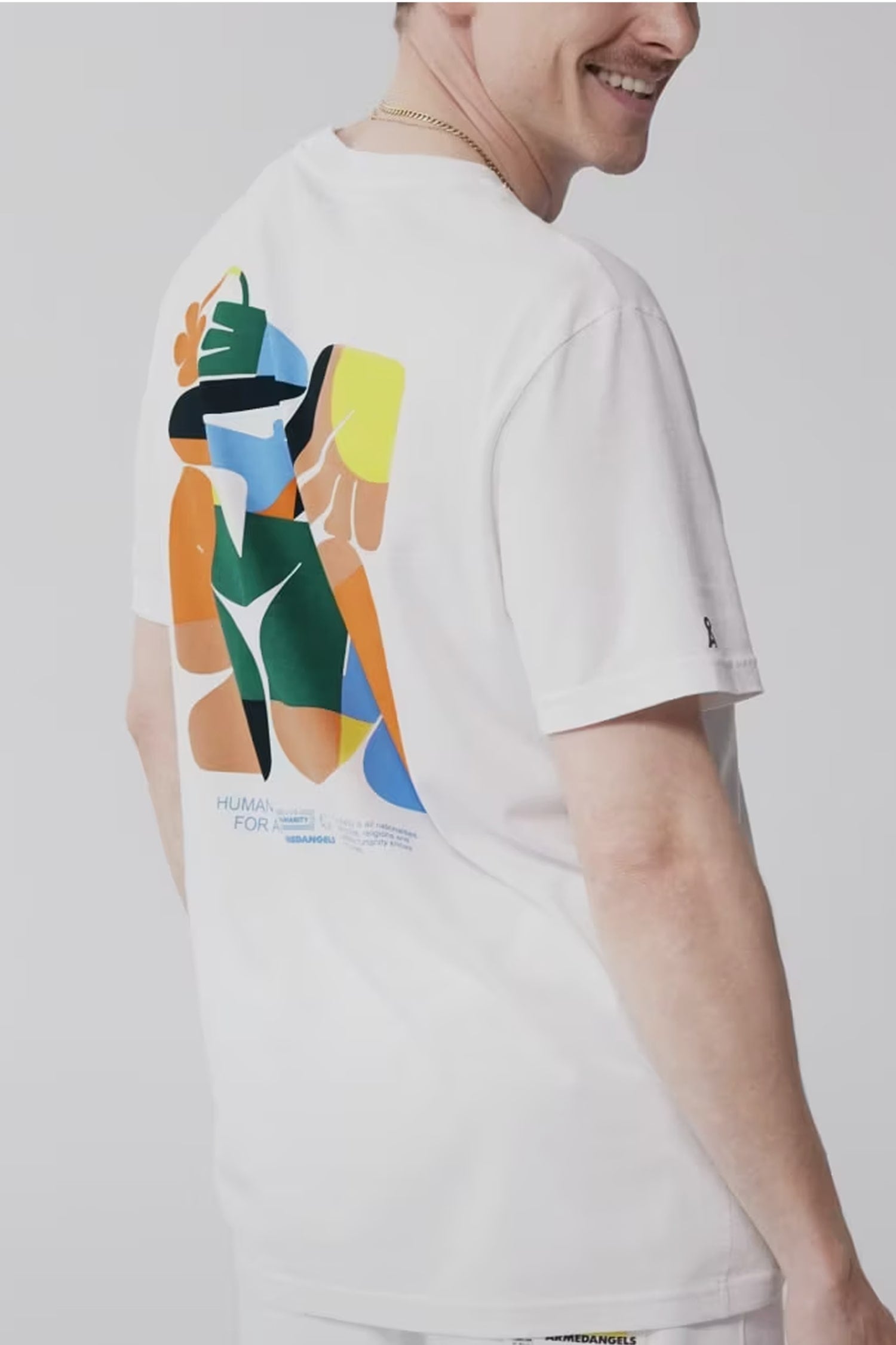 Person trägt weißes T-Shirt mit bunter abstrakter Grafik auf dem Rücken aus der Armedangels-Kollektion ‚Humanity for All‘, vor grauem Hintergrund.
