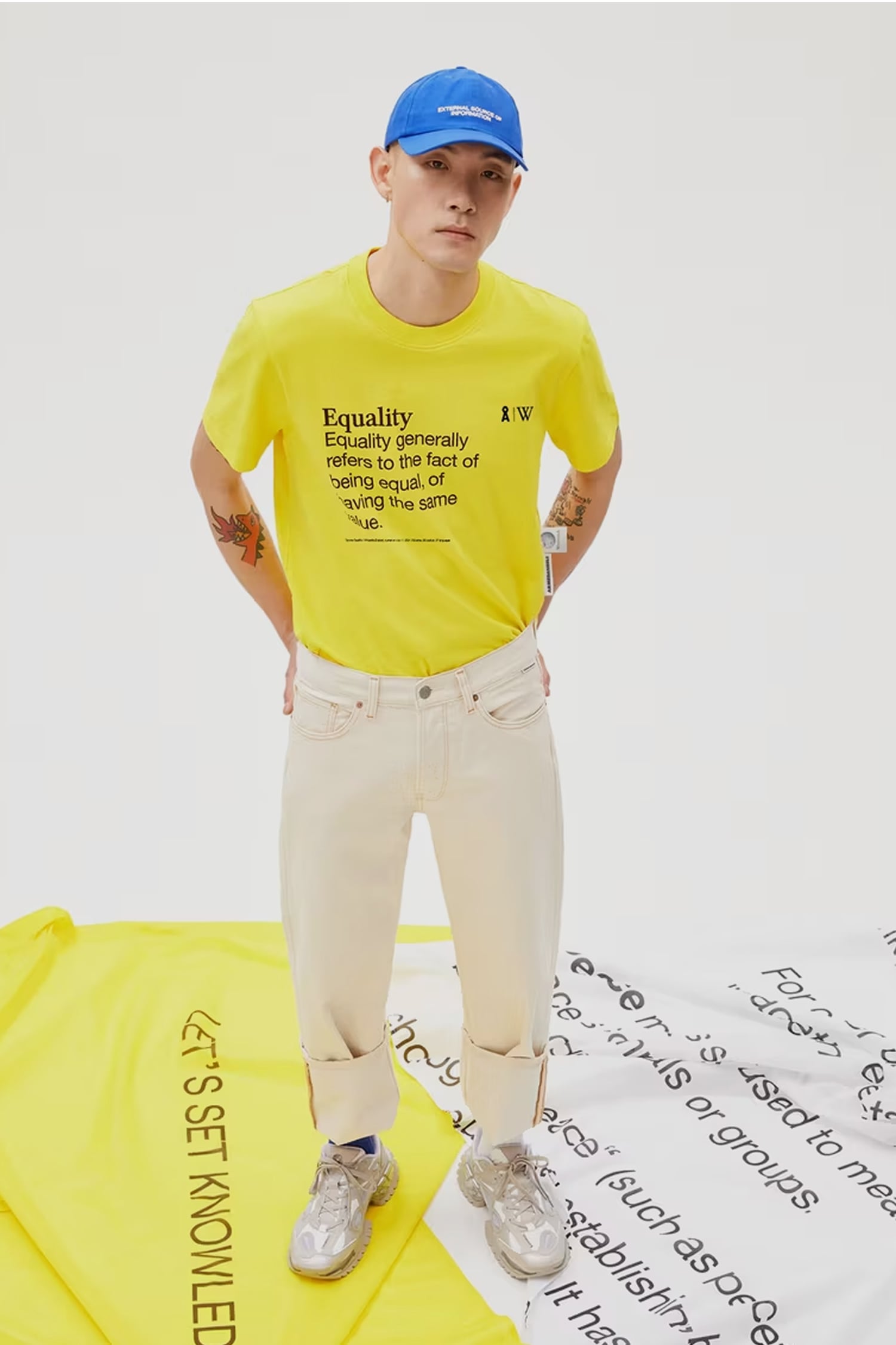 Person trägt gelbes T-Shirt mit Aufdruck ‚Equality‘, cremefarbene Jeans mit umgeschlagenem Saum und blaue Cap, steht auf Stoffbahnen mit Schriftzügen.