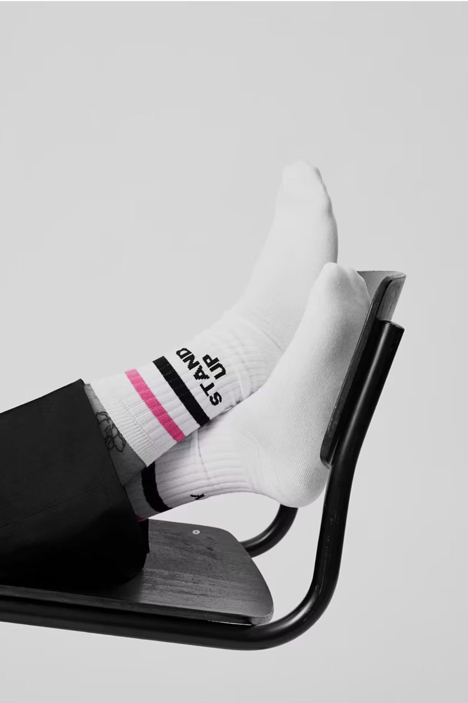 Weiße Socken mit schwarzen und pinken Streifen und dem Schriftzug ‚STAND UP‘, getragen von einer Person, die die Füße auf eine schwarze Metallstuhlstrebe legt.