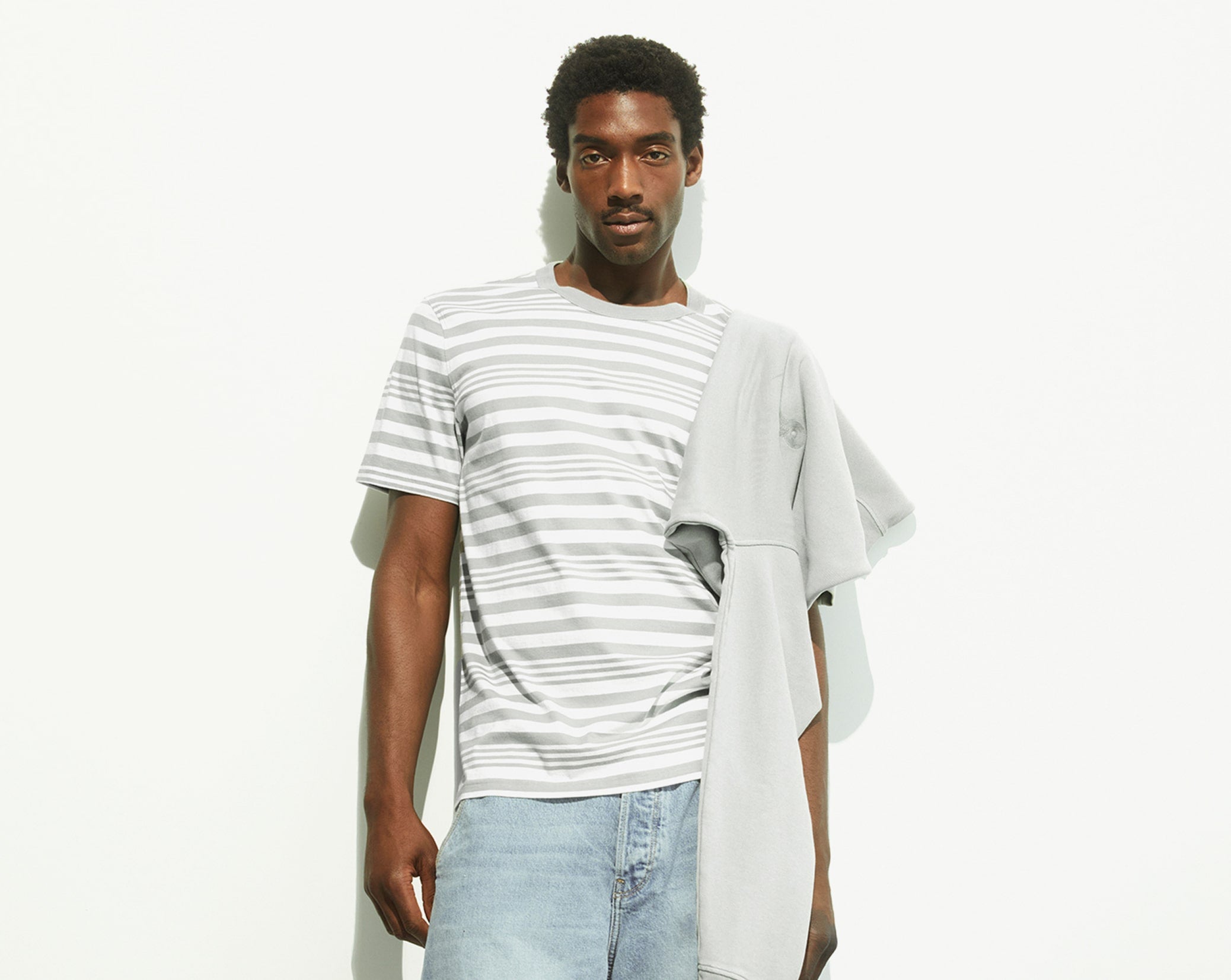 Person trägt grau-weiß gestreiftes T-Shirt, hellblaue Jeans und hat einen grauen Pullover über die Schulter gelegt, stehend vor weißer Wand.