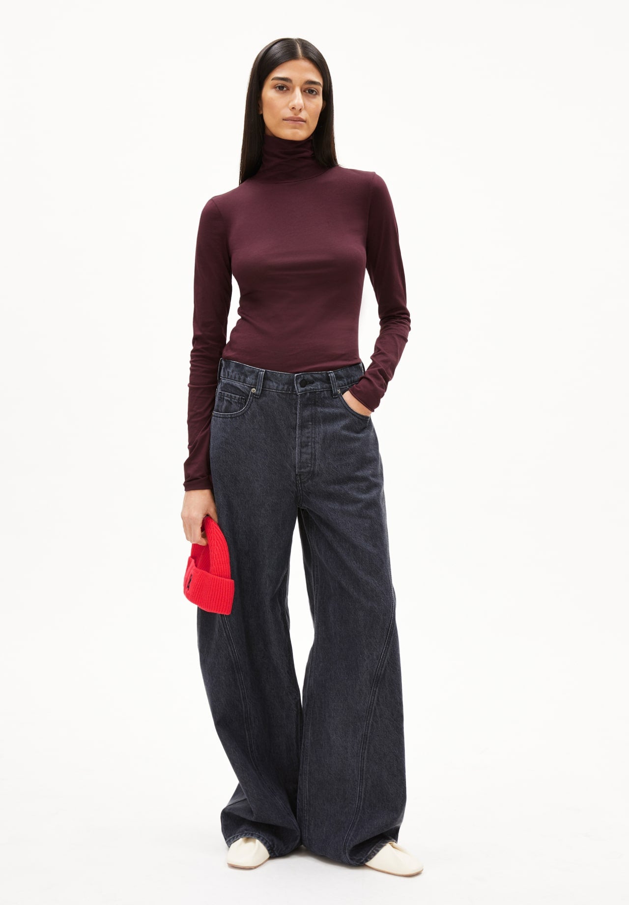 Person trägt ein enges bordeauxfarbenes Rollkragenshirt, eine weite schwarze Jeans und hält eine rote Mütze in der Hand.