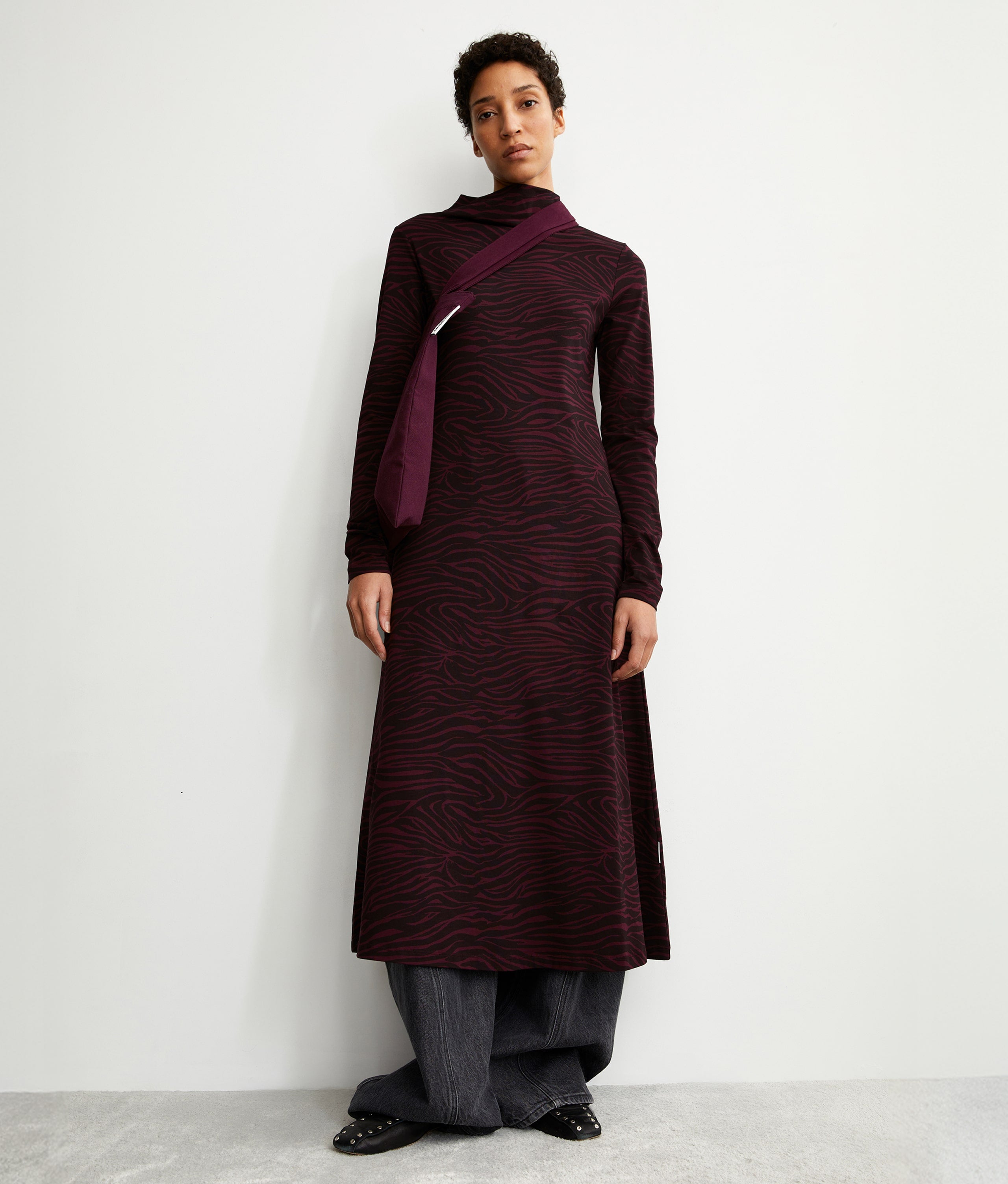 Person trägt ein langes, dunkel gemustertes Kleid mit Rollkragen, eine weite graue Hose darunter und eine diagonale Umhängetasche