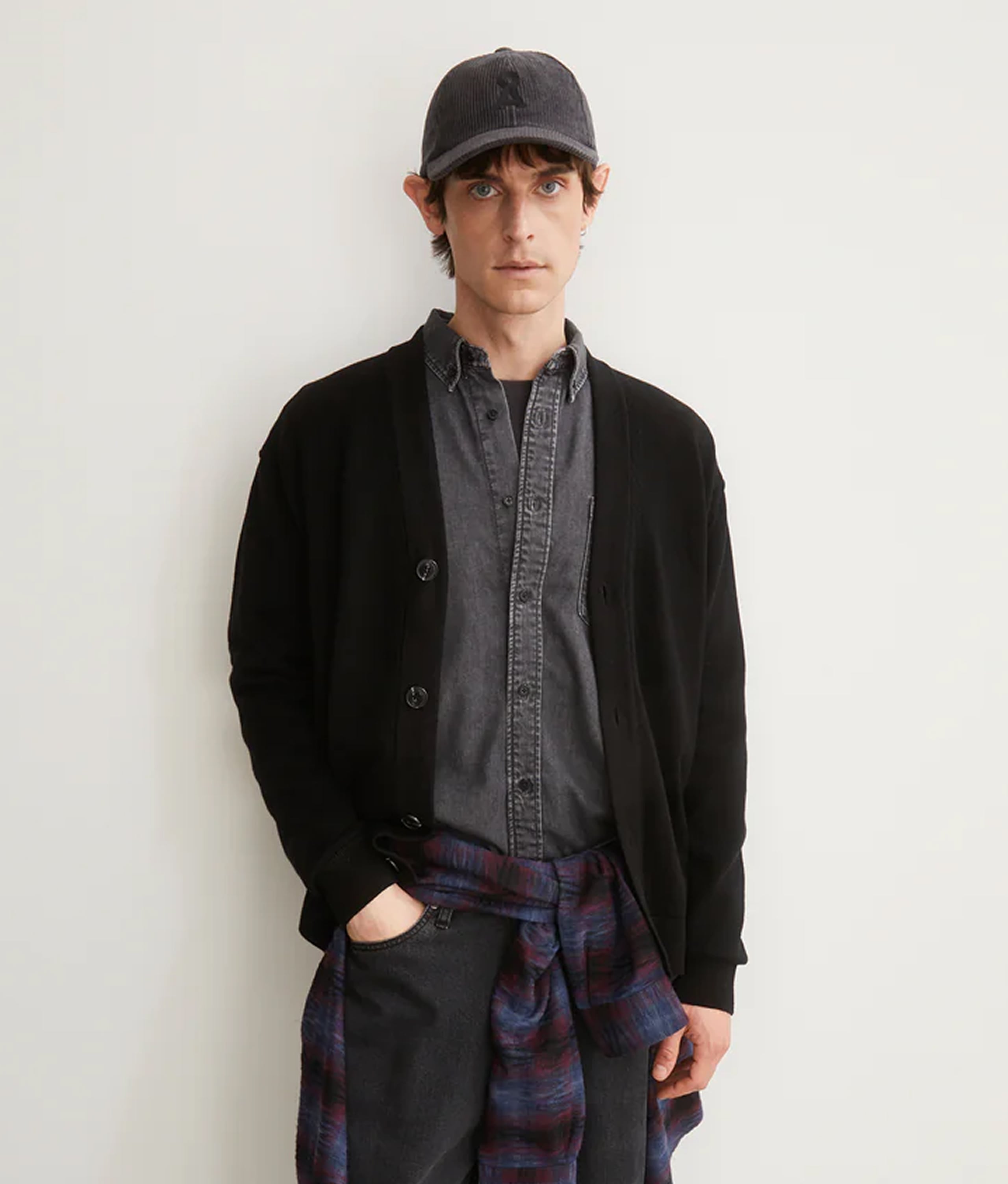 Person trägt ein graues Hemd mit schwarzer Strickjacke, eine graue Kappe und ein kariertes Hemd um die Hüfte gebunden.