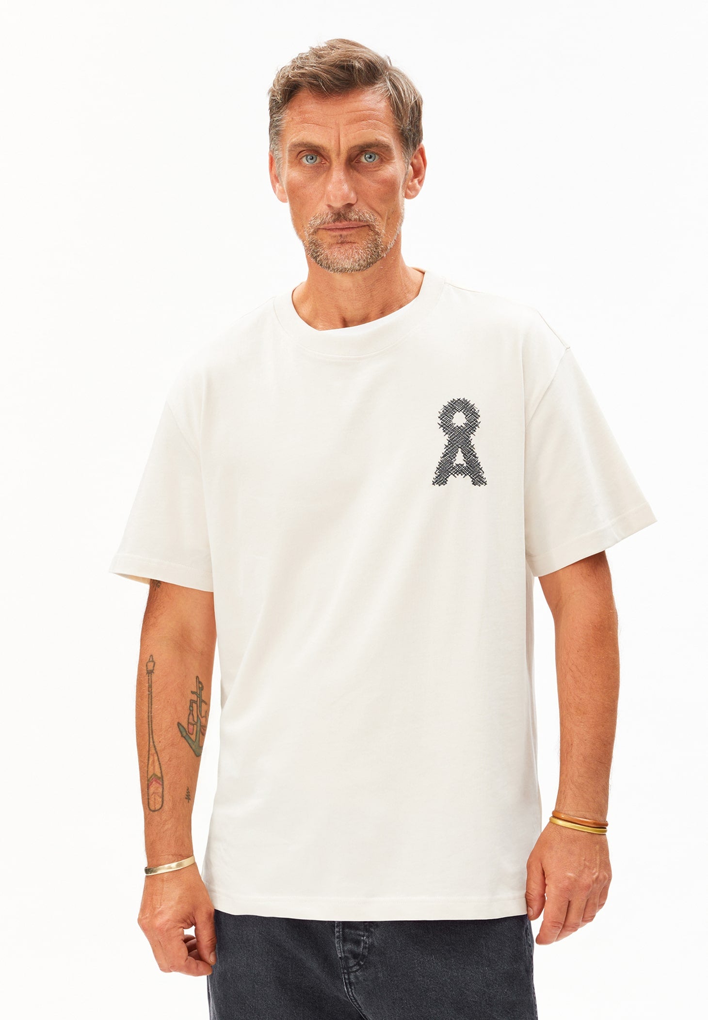 Mann guckt geradeaus und trägt das T-Shirt AADRU Å ZICK ZACK in beige. Dieses hat prominent auf der linken Brust ein gesticktes Å-Logo. Dazu trägt er eine graue Jeans.