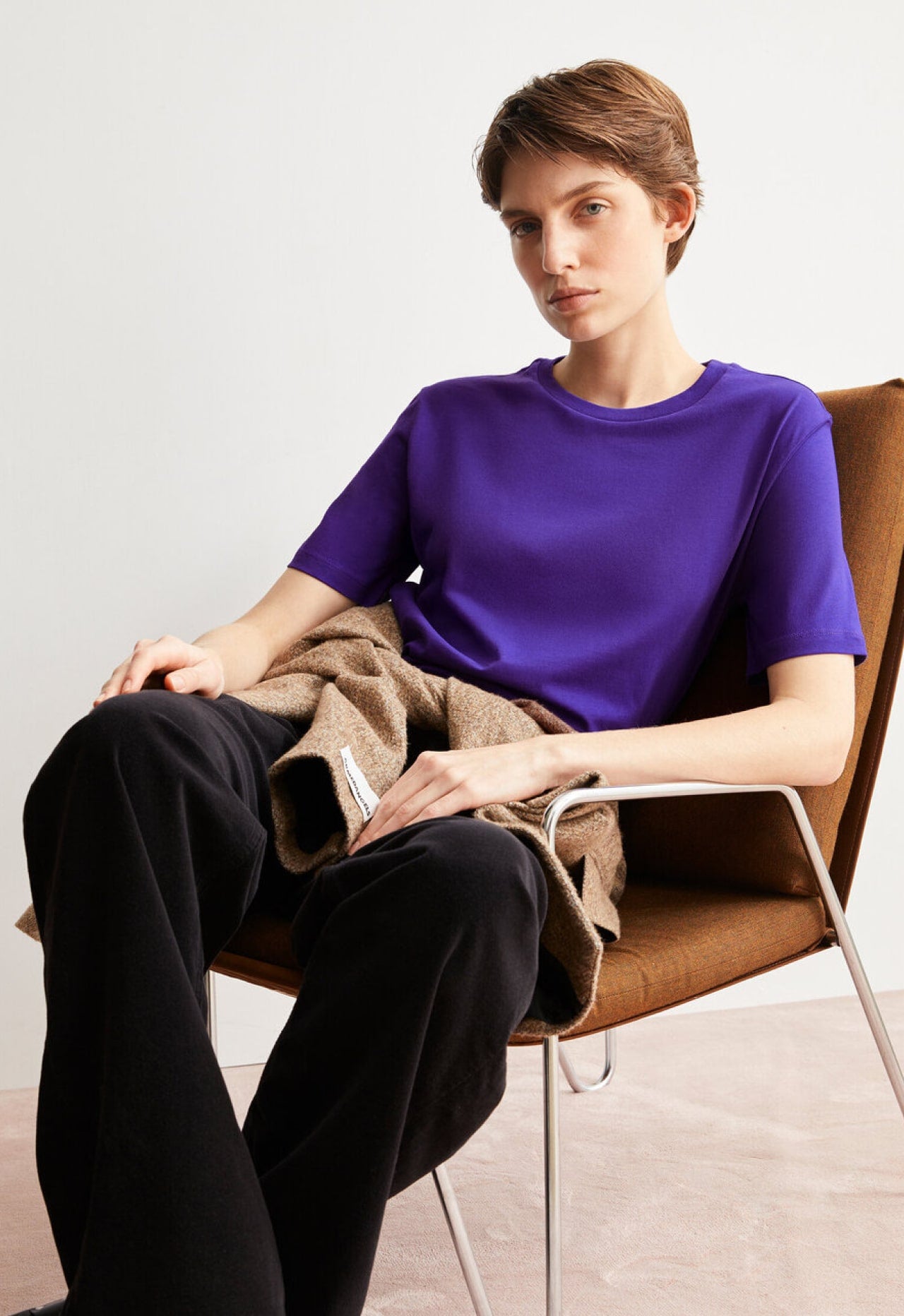 Eine Person sitzt entspannt in einem Stuhl, trägt ein violettes T-Shirt und schwarze Hose und blickt ruhig in die Kamera.