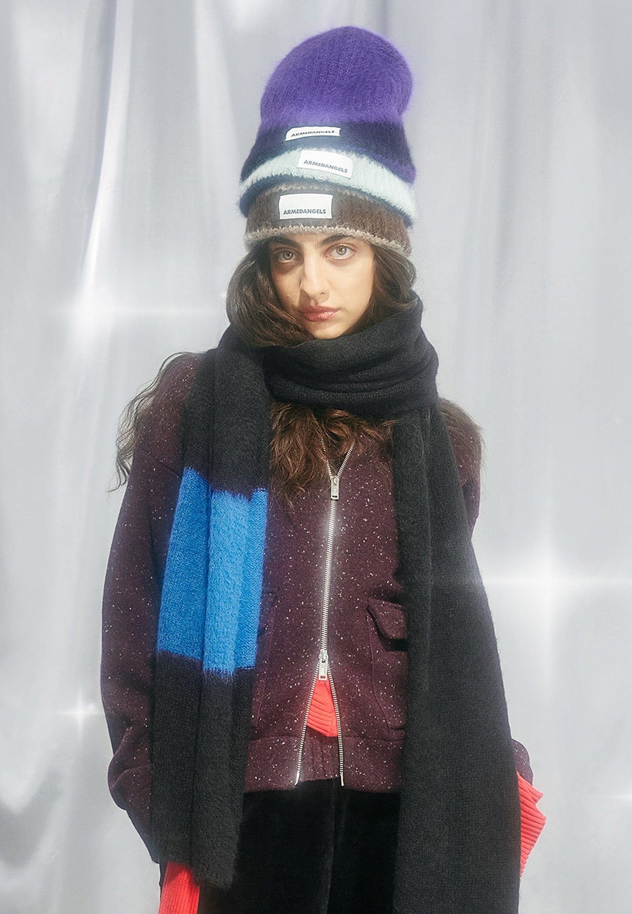 Eine Person trägt mehrere übereinander gestapelte Mützen, einen langen Schal und eine dunkle Zip-Jacke vor hellem Hintergrund.