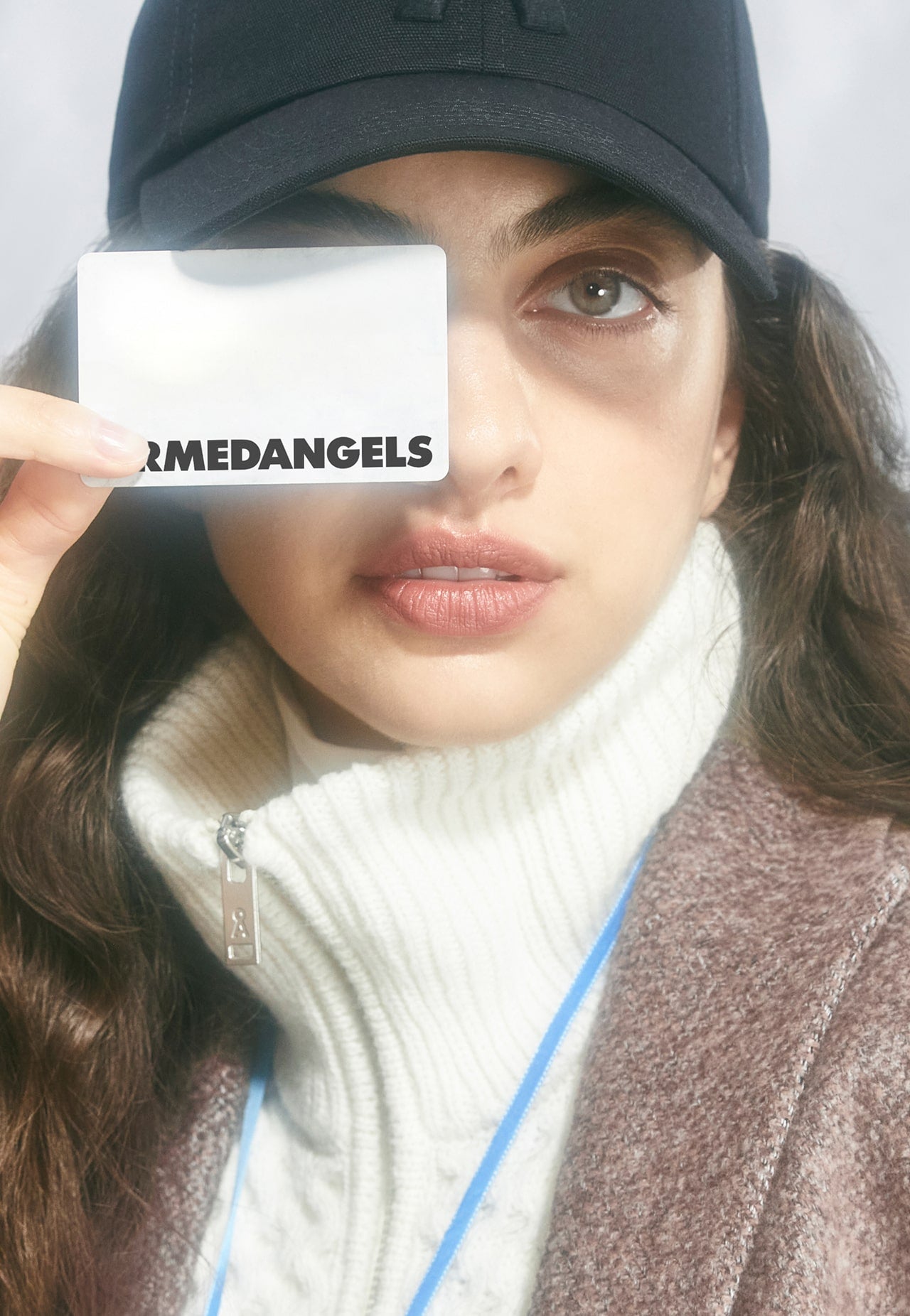 Eine Person trägt eine dunkle Cap und hält eine ARMEDANGELS-Karte vor ein Auge, während sie direkt in die Kamera blickt.