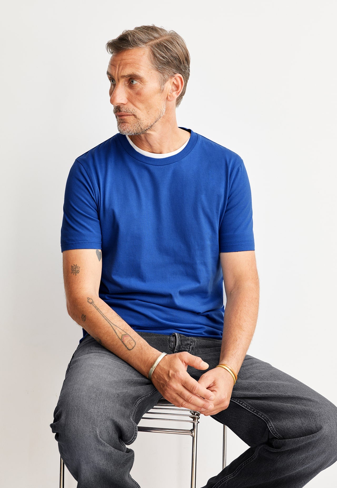 Eine Person sitzt auf einem Hocker, trägt ein blaues T-Shirt und dunkle Jeans und schaut seitlich aus dem Bild.