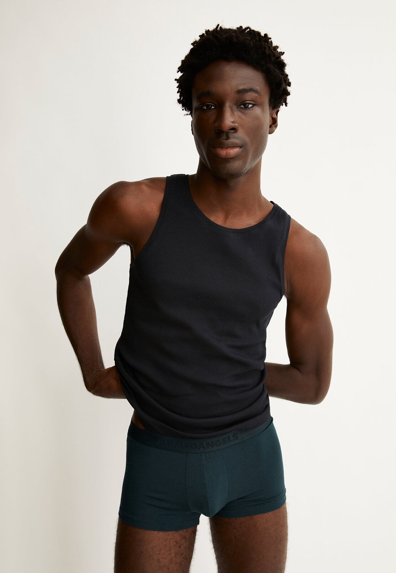 Eine Person trägt ein schwarzes Tanktop und dunkelgrüne Boxershorts und steht vor hellem Hintergrund.