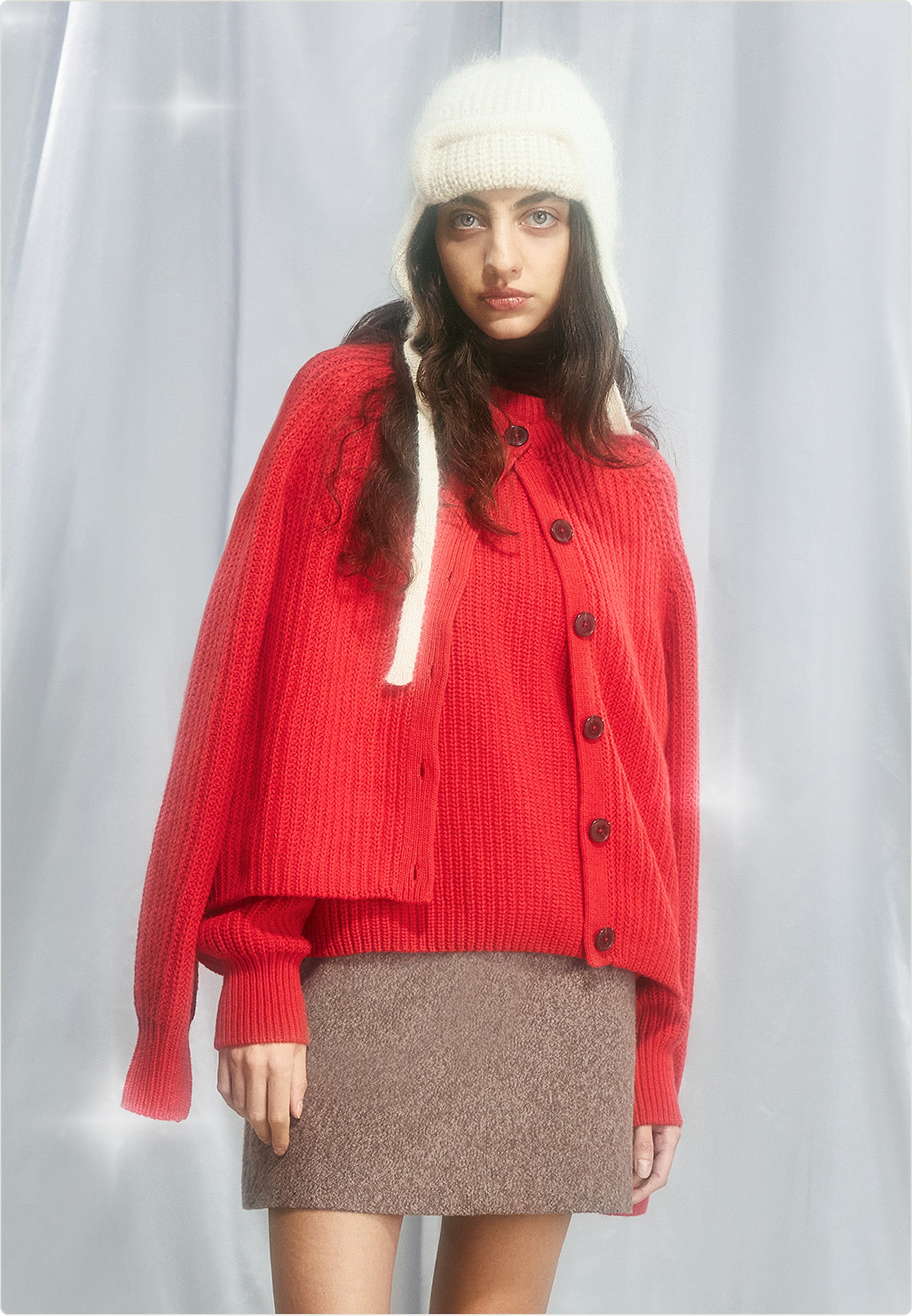 Eine Person trägt eine weiße Strickmütze, einen roten Strickcardigan, einen braunen Minirock und steht vor hellem Vorhang.