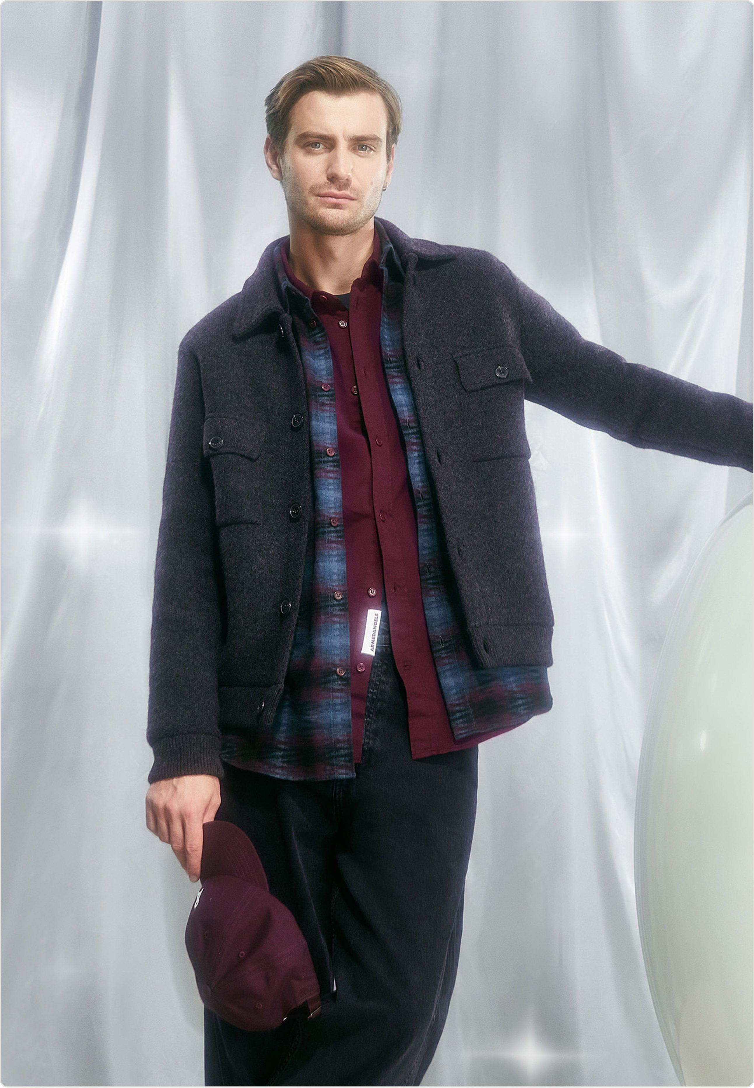 Eine Person trägt eine dunkelgraue Jacke über einem burgunderfarbenen Hemd mit gemusterter Overshirt-Schicht und hält eine passende Cap in der Hand.