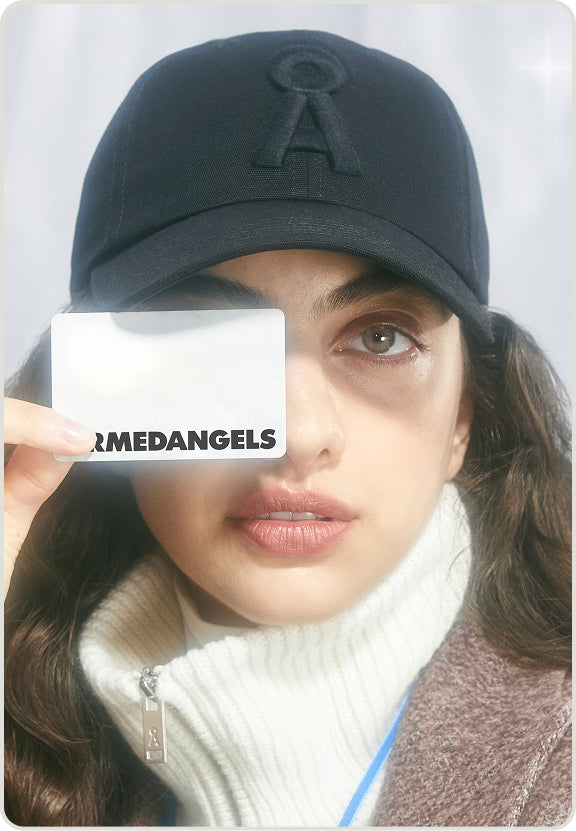 Eine Person trägt eine dunkle Cap und hält eine ARMEDANGELS-Karte vor ein Auge, während sie direkt in die Kamera blickt.