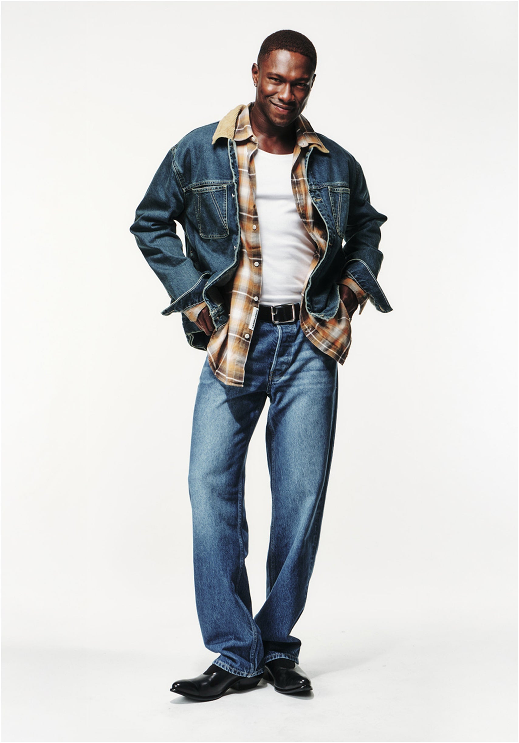 Person steht vor hellem Hintergrund, trägt Jeansjacke mit Cordkragen, kariertes Hemd, weißes Tanktop, Jeans und schwarze Stiefel, mit entspanntem, selbstbewusstem Ausdruck.