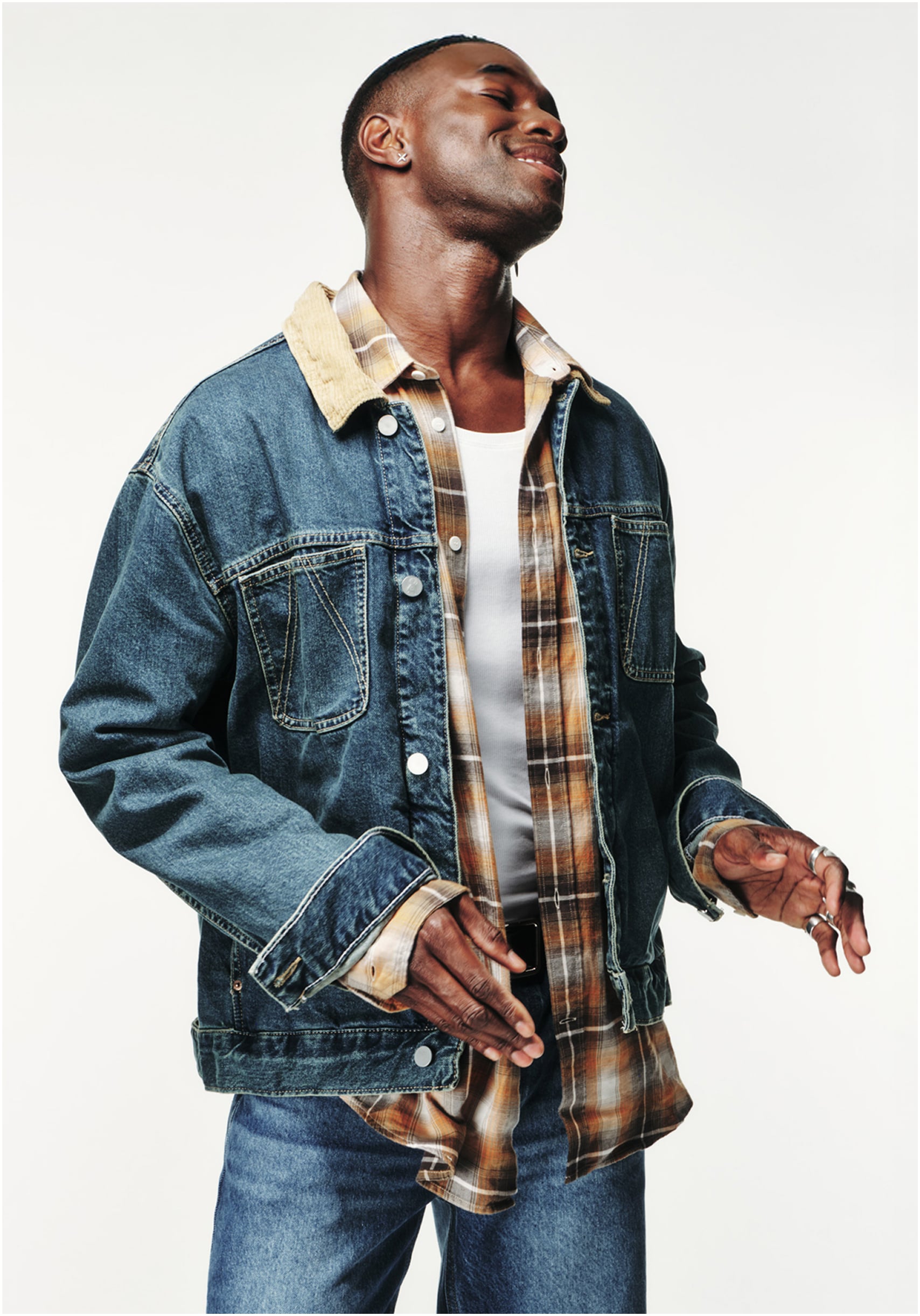 Person vor hellem Hintergrund, trägt Jeansjacke mit Cordkragen über kariertem Hemd und weißem Tanktop, lächelt mit geschlossenen Augen und bewegt sich entspannt.