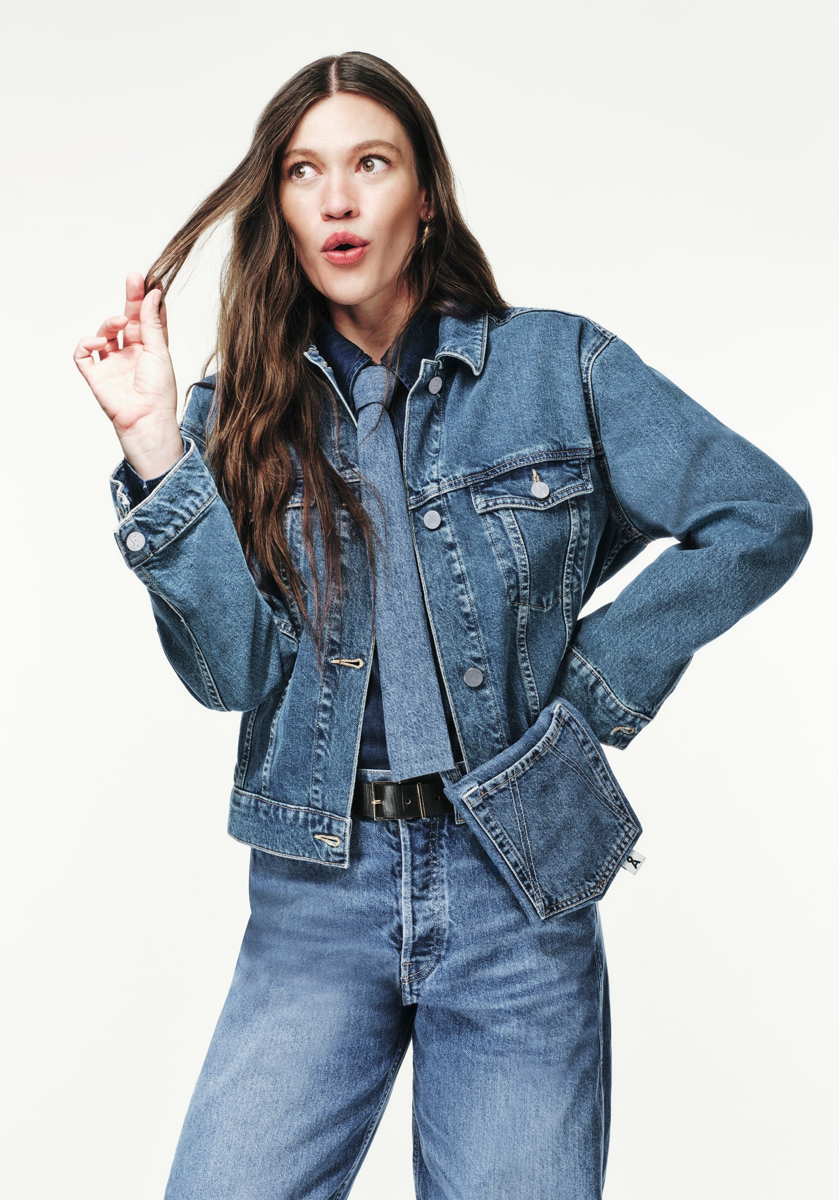 Person steht vor hellem Hintergrund, trägt Jeansjacke, Jeanshemd, Krawatte und Jeanshose, spielt mit einer Haarsträhne und blickt überrascht oder nachdenklich zur Seite.