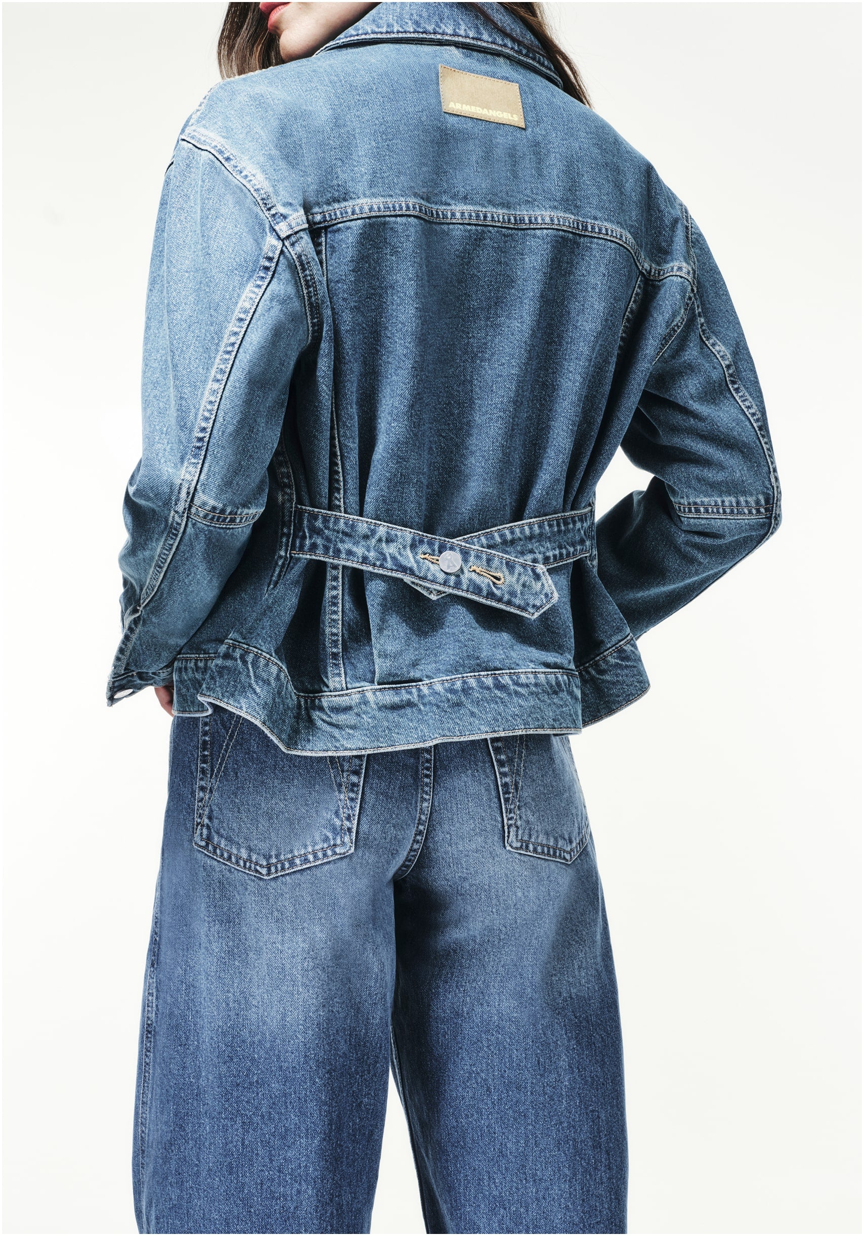 Person im Rückenansicht trägt eine locker sitzende Jeansjacke mit Gürtel-Detail und dazu passende Jeans, vor hellem Hintergrund.