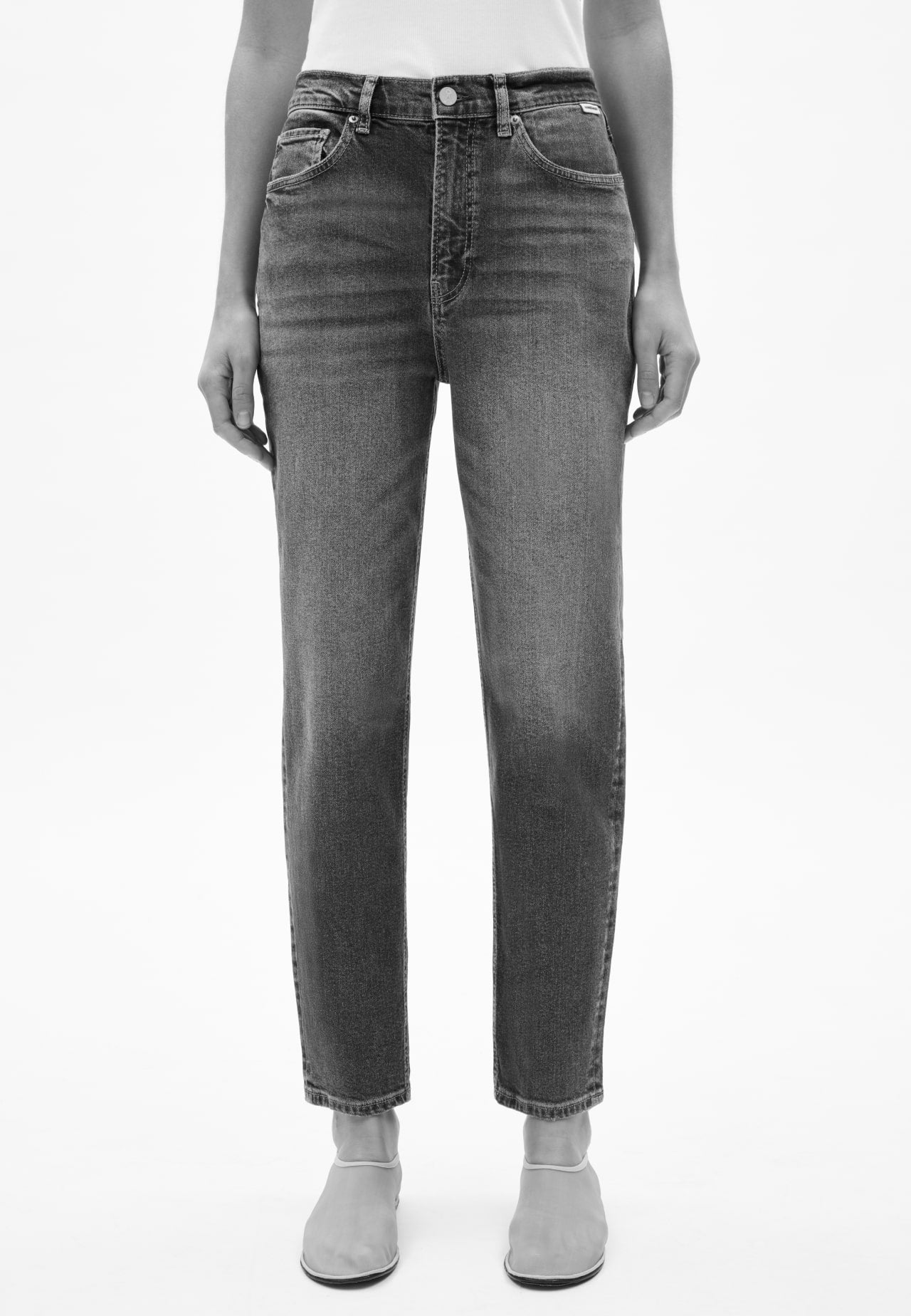 Vorderansicht einer Person mit schwarzer Jeans in geradem Schnitt, kombiniert mit weißem Oberteil und hellen Schuhen, vor hellem Hintergrund.