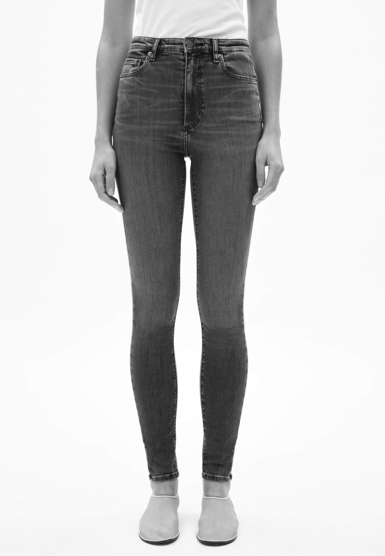 Vorderansicht einer Person mit schwarzer Jeans in geradem Schnitt, kombiniert mit weißem Oberteil und hellen Schuhen, vor hellem Hintergrund.