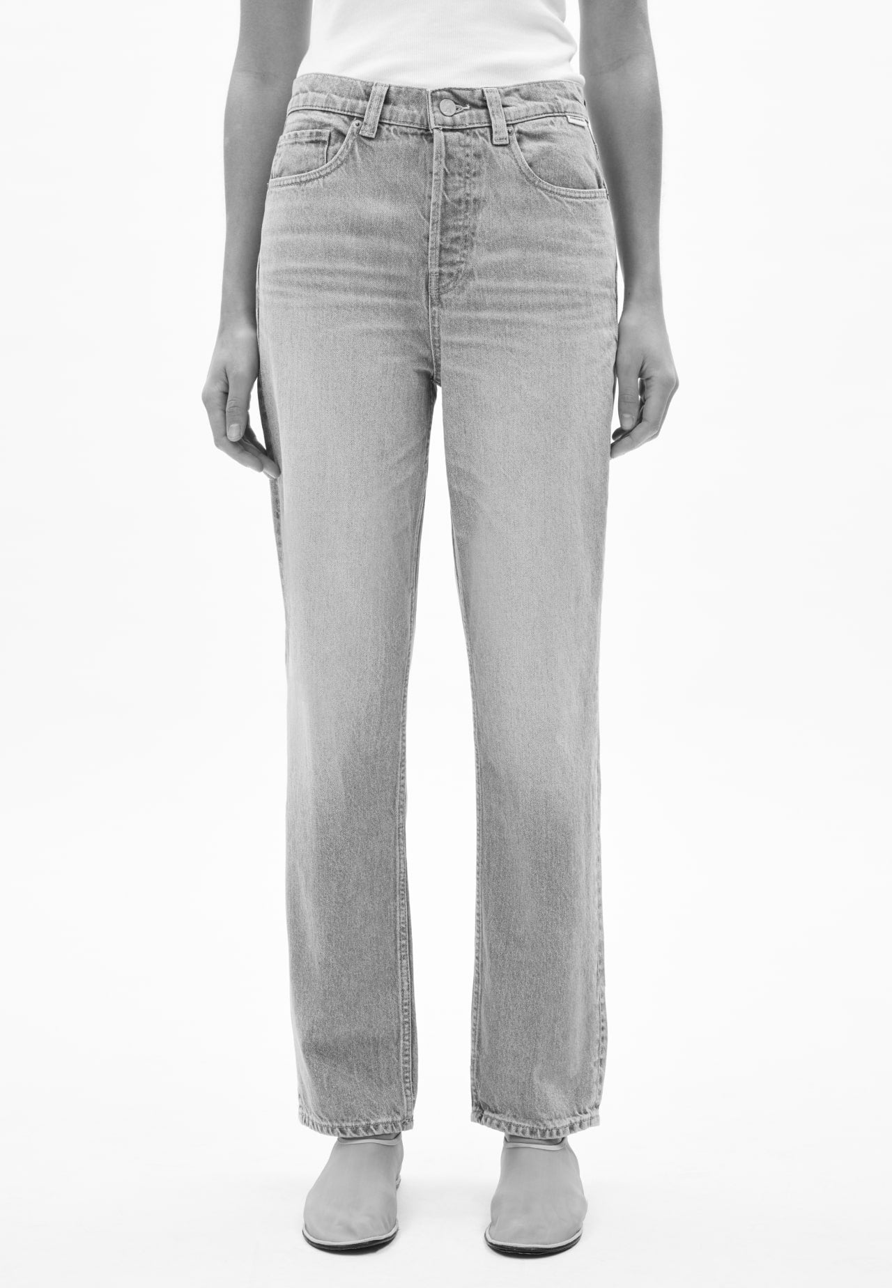 Vorderansicht einer Person mit blauer Jeans in geradem Schnitt, kombiniert mit weißem Oberteil und hellen Schuhen, vor hellem Hintergrund.