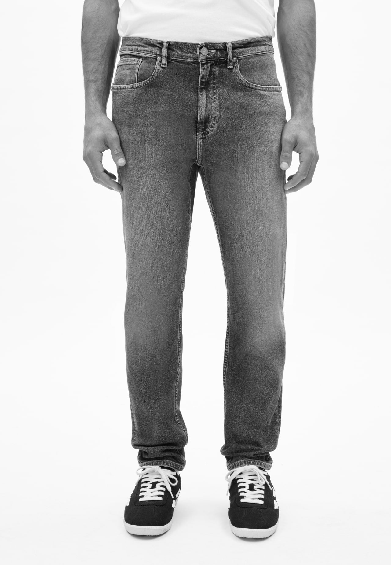 Person trägt eine schmal geschnittene Jeans in dunklem Grau und schwarze Sneaker mit weißen Schnürsenkeln, vor neutralem Hintergrund.