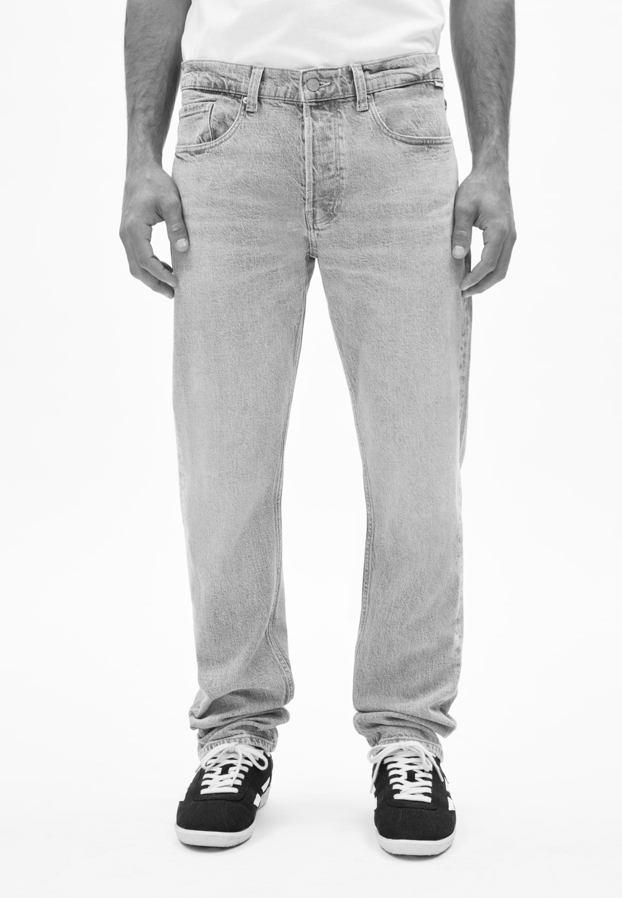 Person trägt eine Jeans in dunklem Grau und schwarze Sneaker mit weißen Schnürsenkeln, vor neutralem Hintergrund.