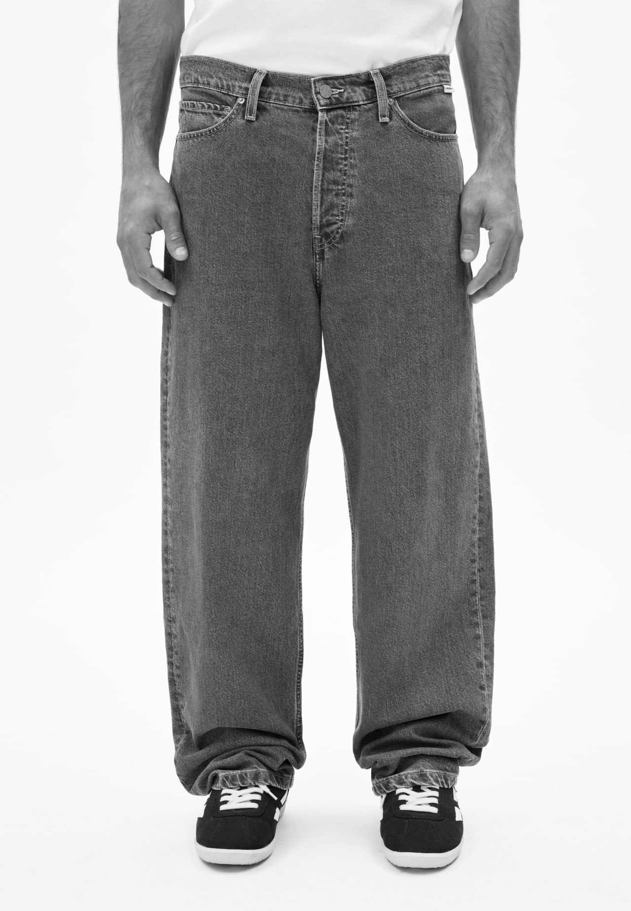 Person trägt eine Jeans in dunklem Grau und schwarze Sneaker mit weißen Schnürsenkeln, vor neutralem Hintergrund.