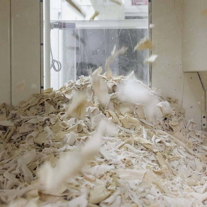 Haufen aus hellen Textilresten, die in einer Maschine zerkleinert und aufgewirbelt werden.