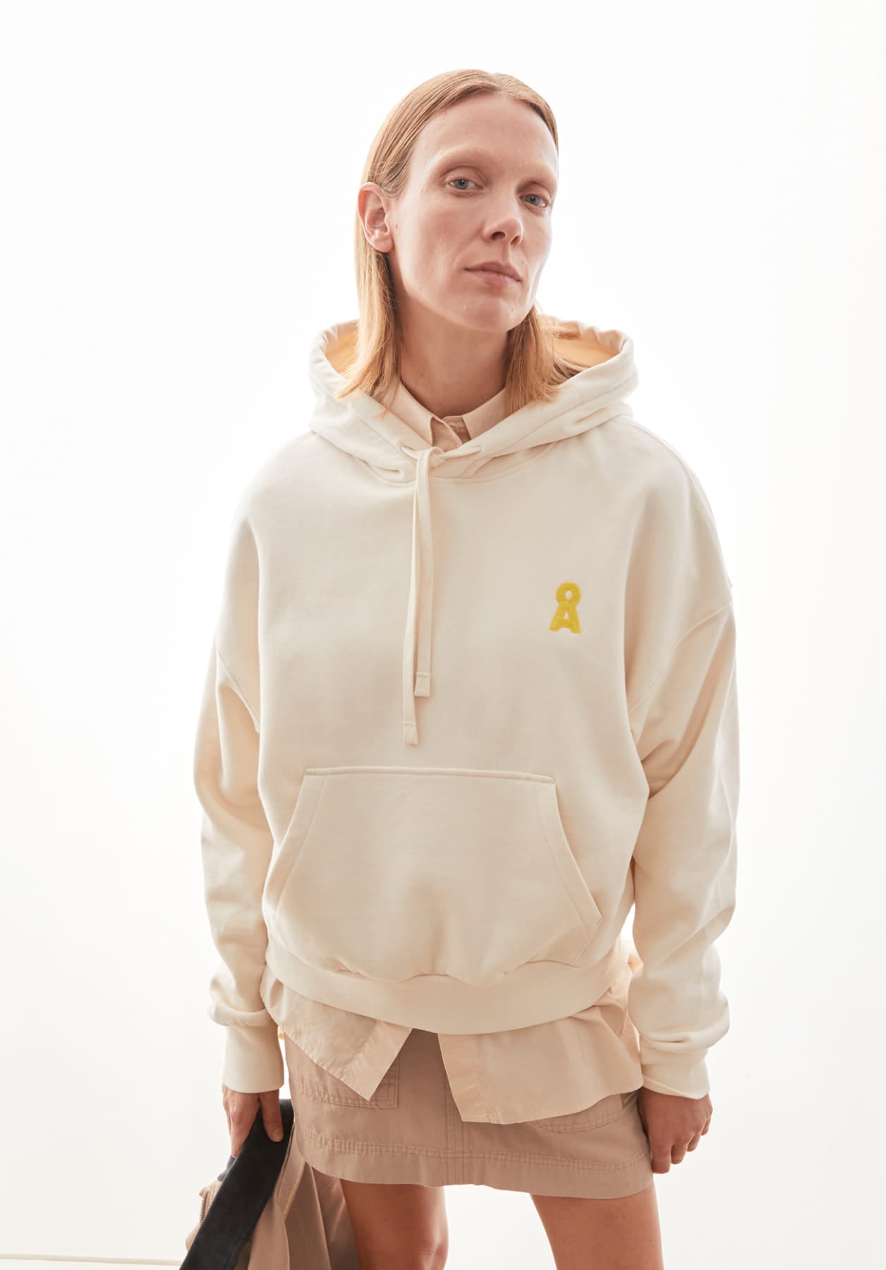 Ein Model steht vor hellem Hintergrund und trägt einen cremefarbenen Hoodie mit Logo, kombiniert mit beigem Rock und lässigem Layering.