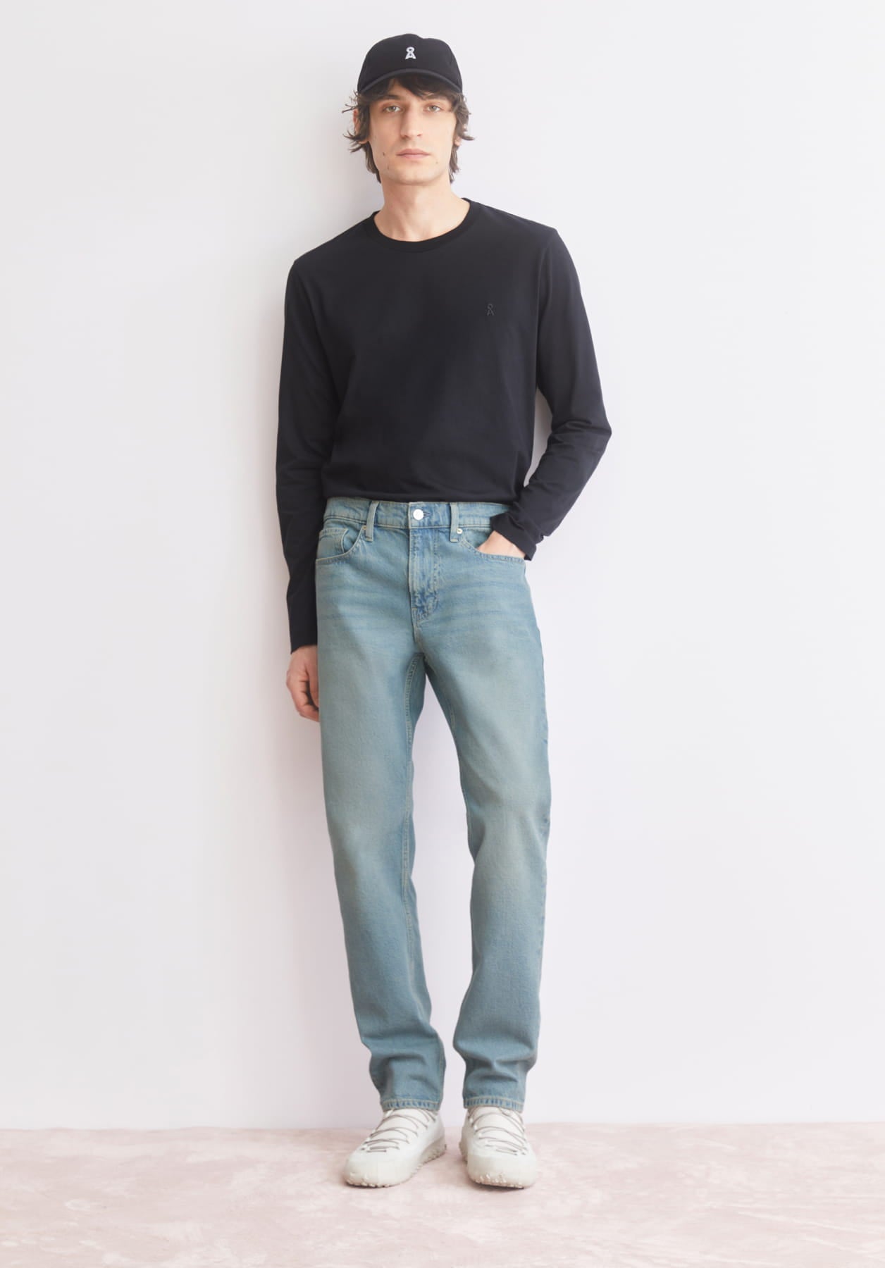 Ein Model steht vor einer hellen Wand und trägt ein schwarzes Longsleeve, helle Jeans, weiße Sneaker und eine schwarze Cap.