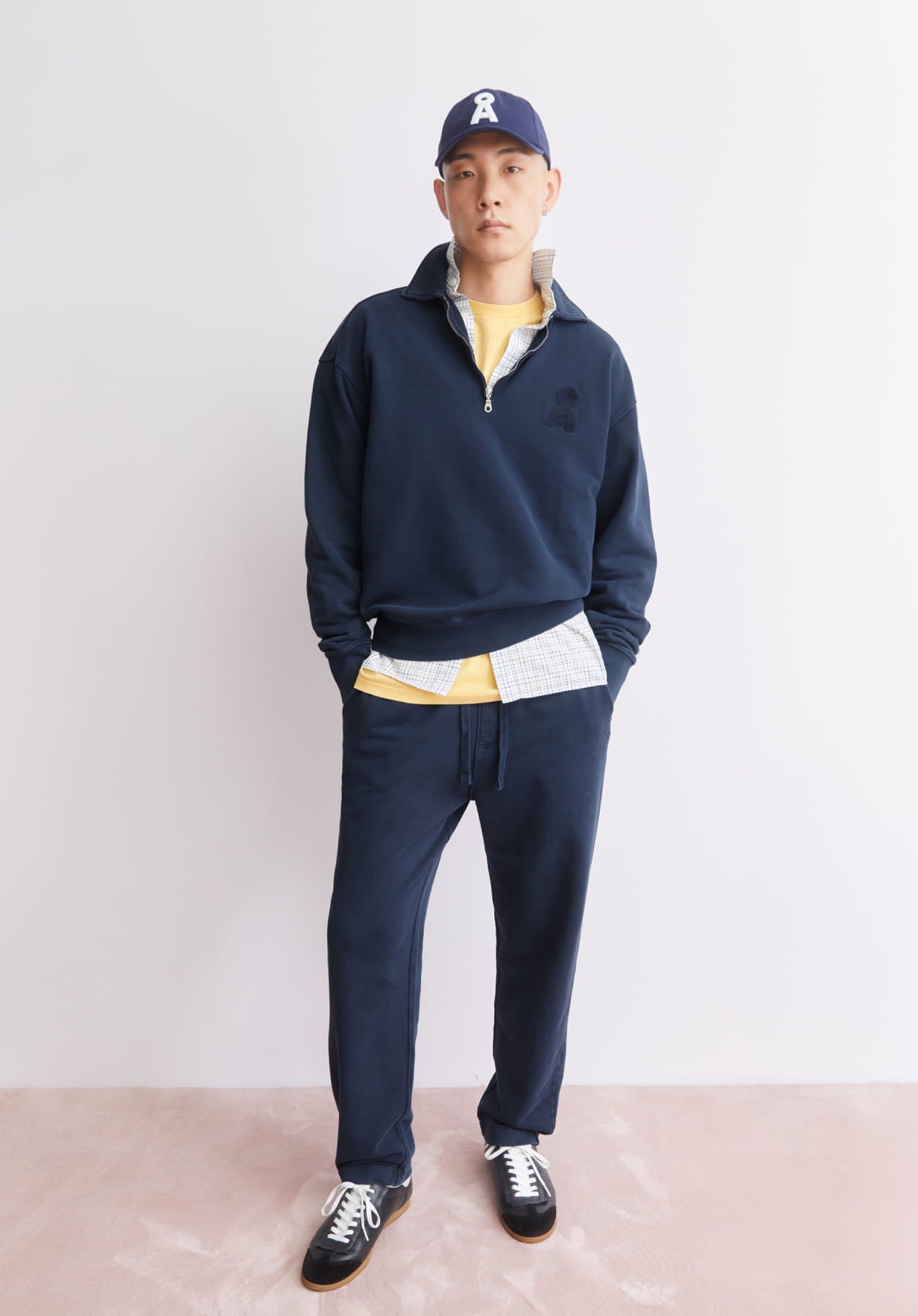 Ein Model steht vor heller Wand und trägt einen dunkelblauen Half-Zip-Pullover mit passender Jogpants, gelbem Shirt, Cap und Sneakern.