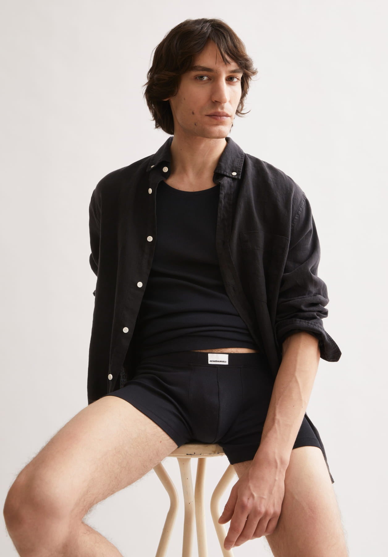 Ein Model sitzt vor hellem Hintergrund auf einem Hocker und trägt ein schwarzes Tanktop, offenes Hemd und schwarze Shorts.