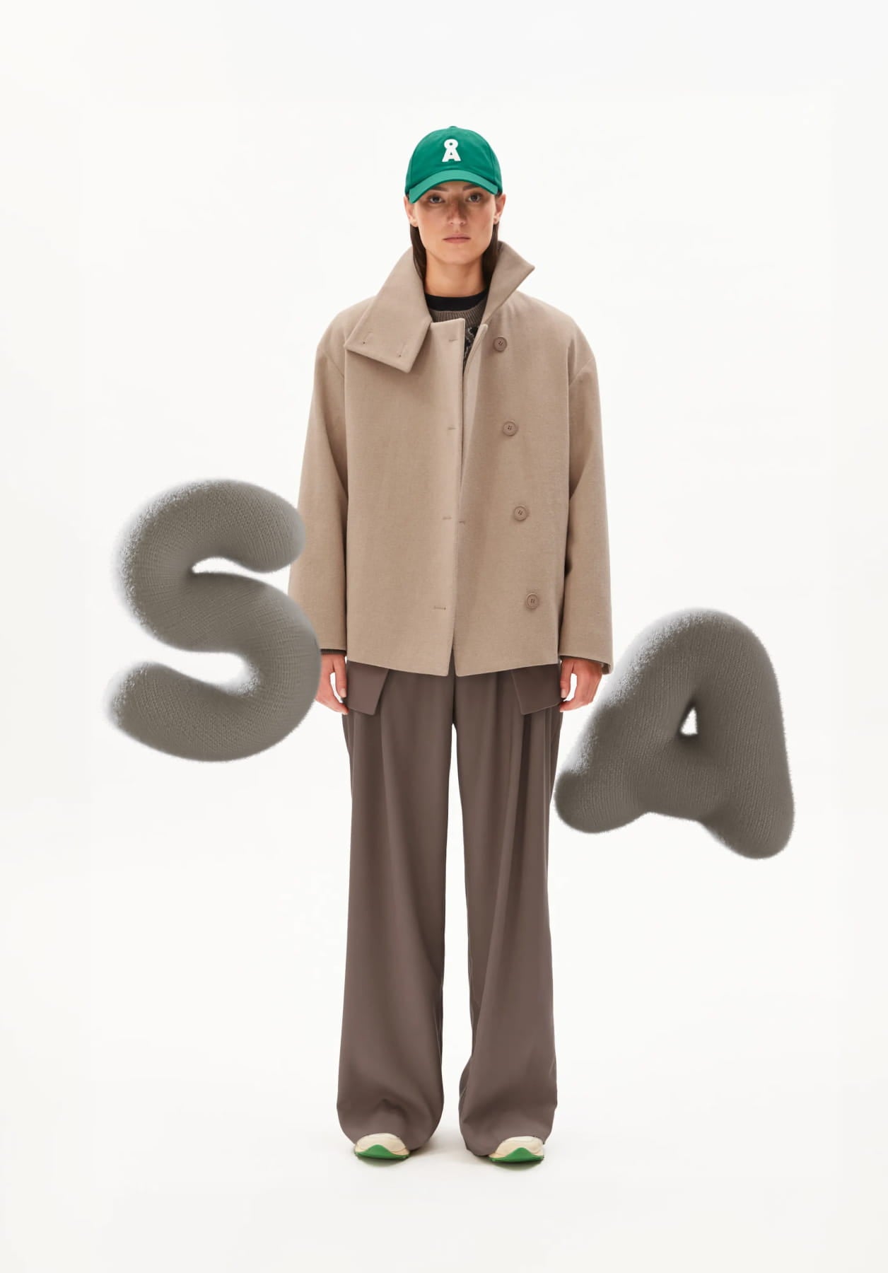 Ein Model trägt vor hellem Hintergrund einen beigen Kurzmantel, weite Stoffhose und eine grüne Cap, begleitet von grafischen, abstrakten Buchstaben.