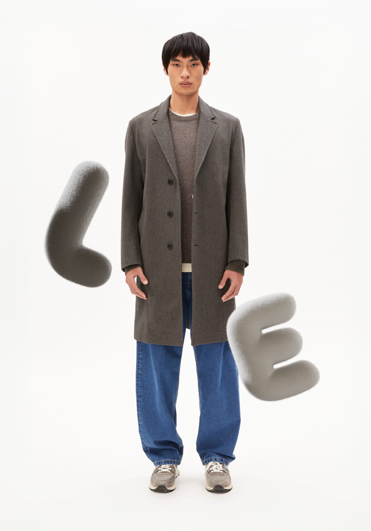 Ein Model steht vor hellem Hintergrund und trägt einen grauen Mantel mit Strickpullover, blauer Jeans und Sneakern, ergänzt durch grafische, abstrakte Elemente.