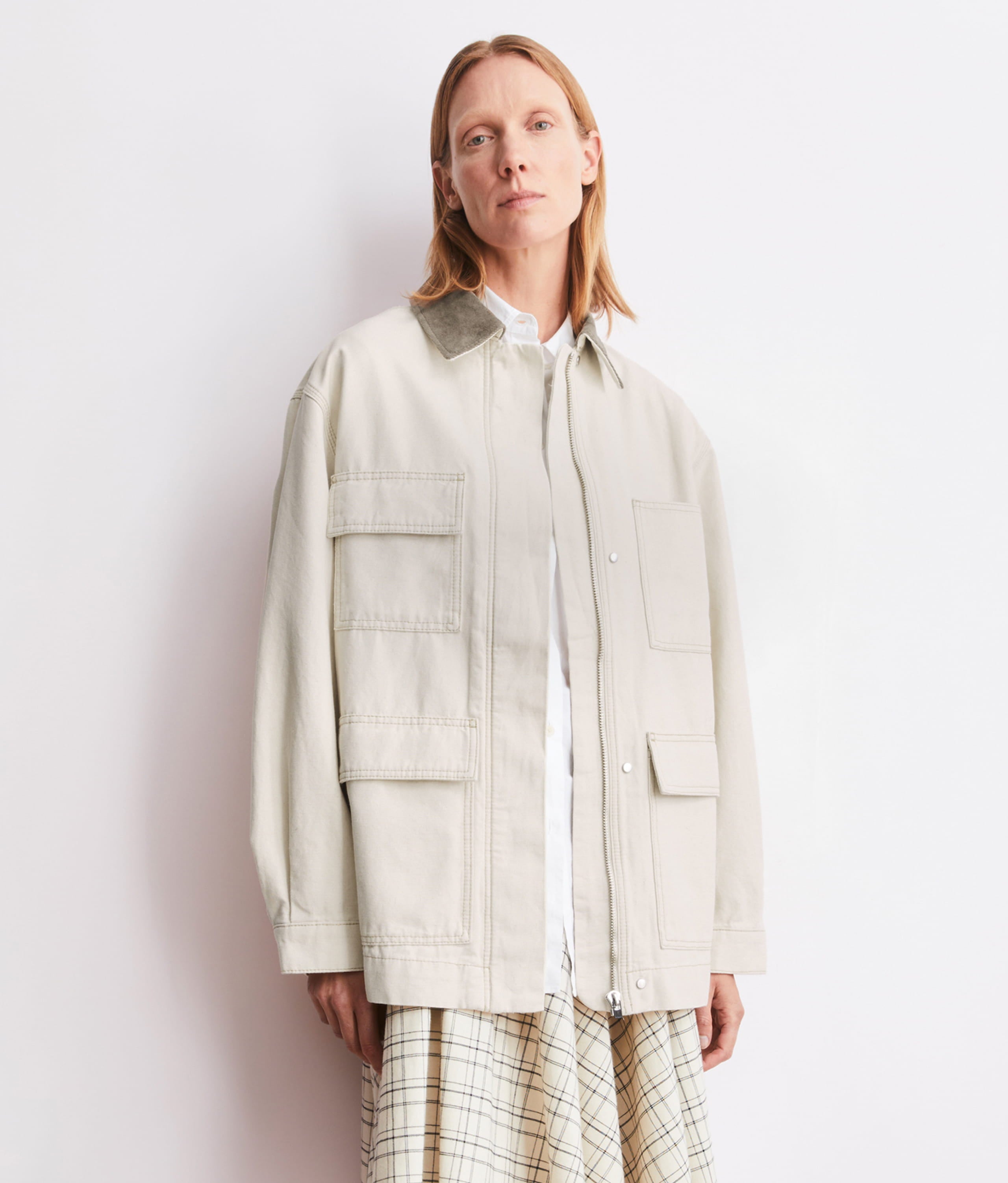 Ein Model steht vor hellem Hintergrund und trägt eine helle Utility-Jacke über weißem Hemd, kombiniert mit gemustertem Rock.