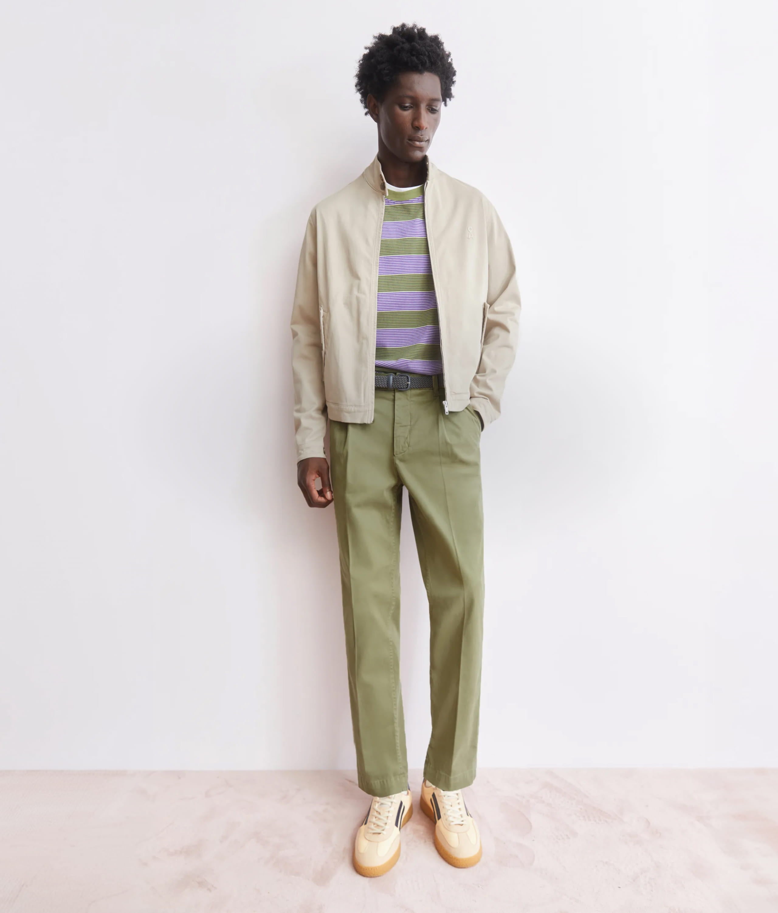 Ein Model steht vor hellem Hintergrund und trägt eine helle Jacke über gestreiftem Shirt, olivgrüne Hose und helle Sneaker.