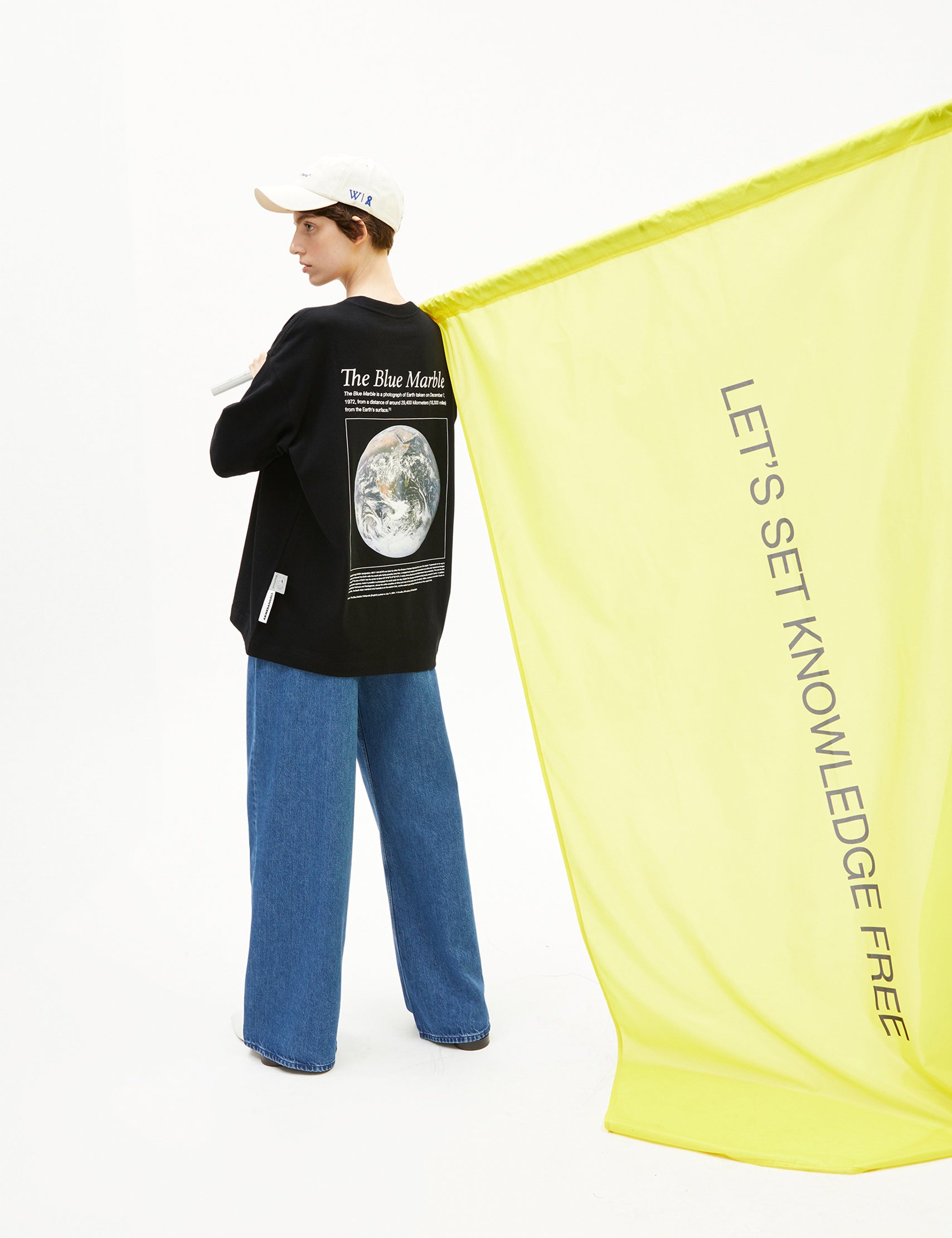 Model trägt schwarze Sweatshirt mit Earth-Print ‚The Blue Marble‘ und hält gelbes Banner mit Schriftzug ‚Let’s set knowledge free‘.