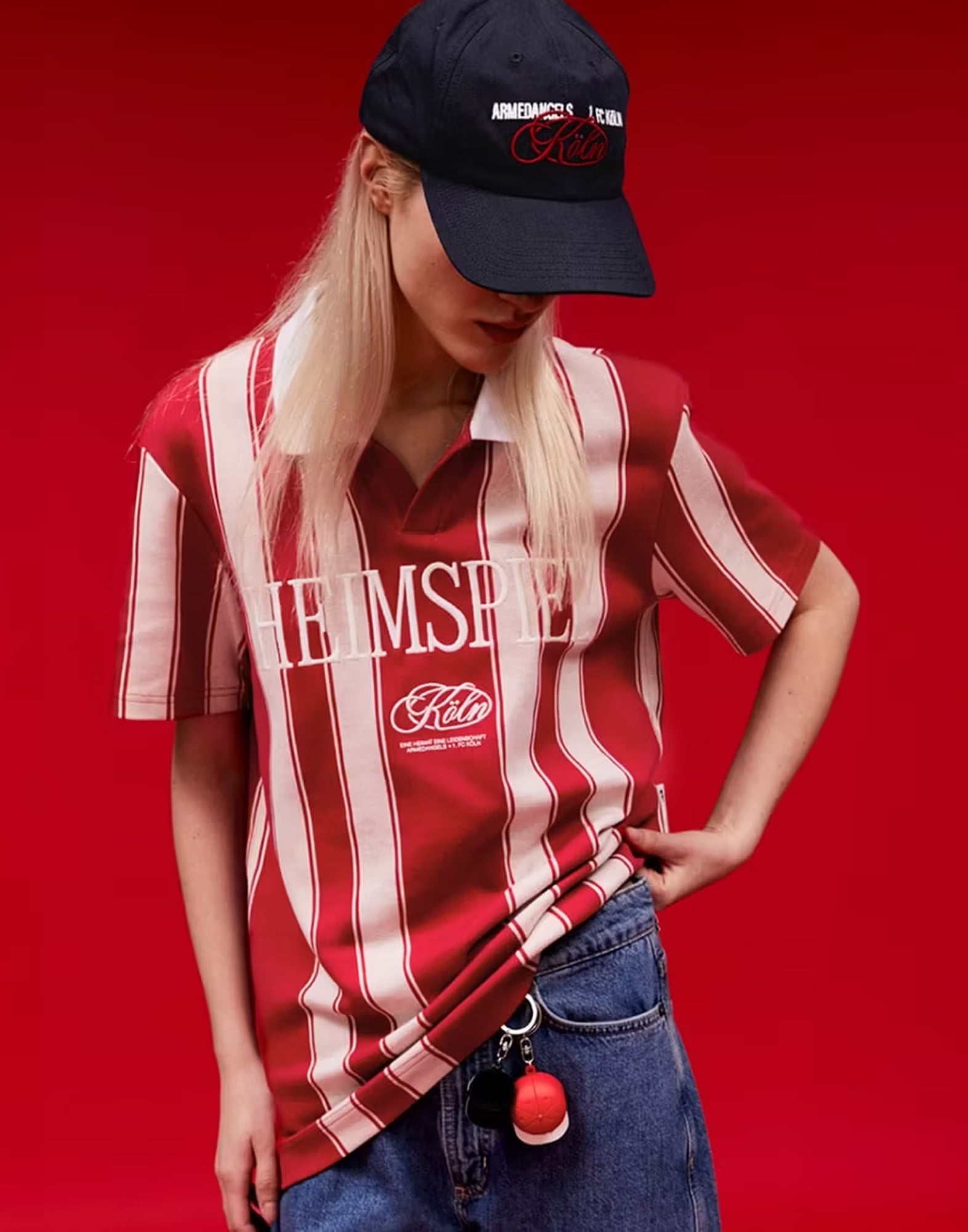 „Person trägt rot-weiß gestreiftes Shirt mit Aufdruck ‚HEIMSPIEL Köln‘ und schwarze Cap mit weiß-roter Schrift, kombiniert mit blauer Jeans, vor rotem Hintergrund.