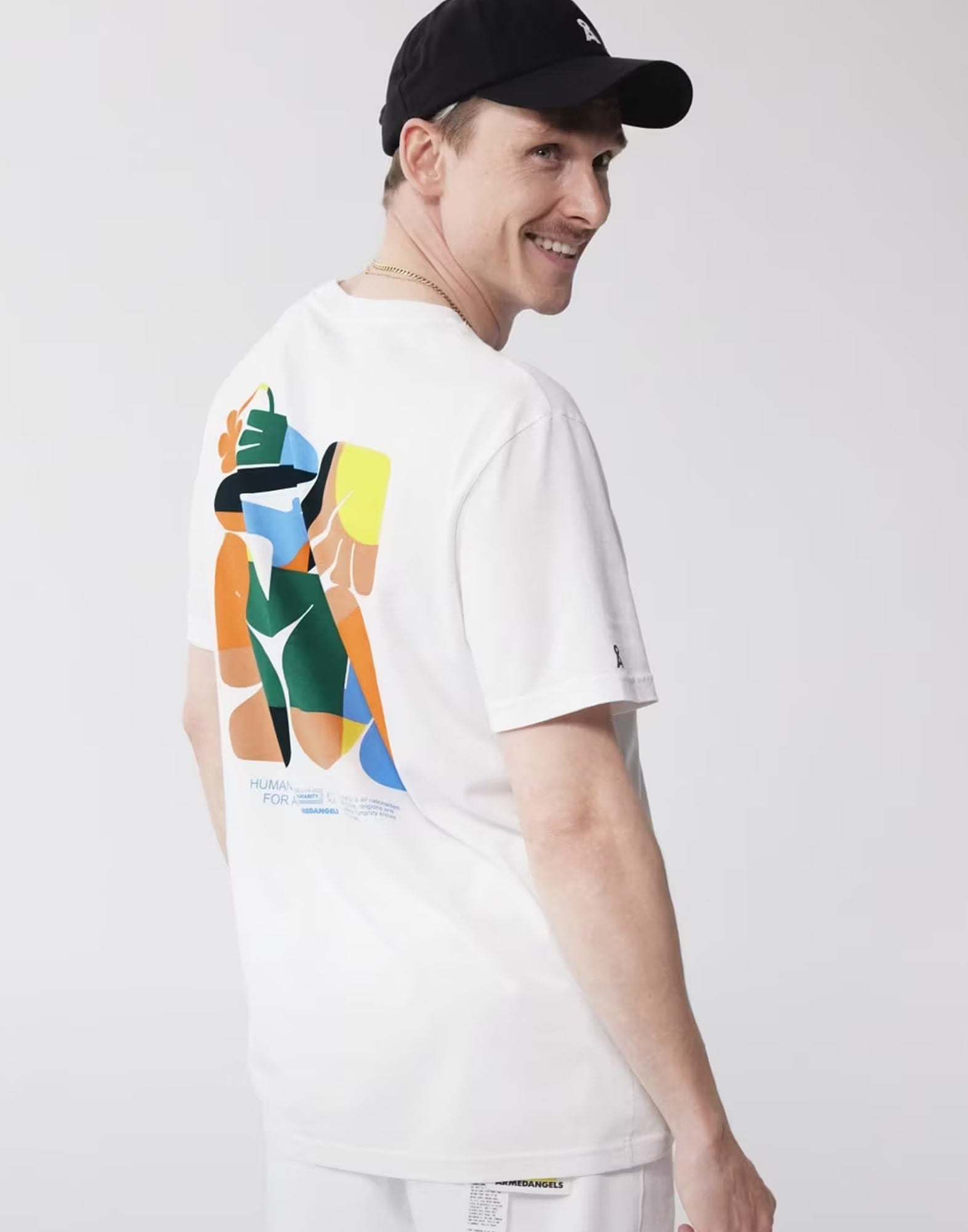 Person trägt weißes T-Shirt mit bunter abstrakter Grafik auf dem Rücken aus der Armedangels-Kollektion ‚Humanity for All‘, vor grauem Hintergrund.
