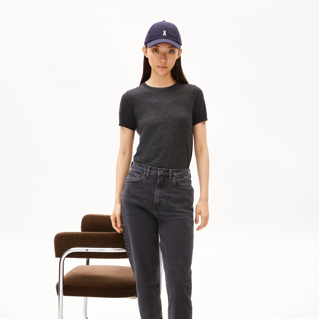 Person trägt dunkles T-Shirt, graue Jeans und eine dunkelblaue Cap, steht vor einem braunen Stuhl auf hellem Hintergrund.