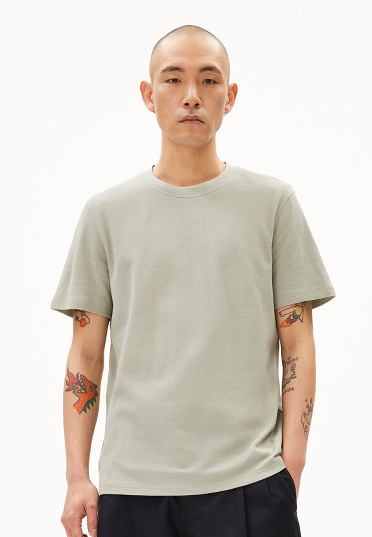 Person trägt hellgraues relaxed fitted T-Shirt aus Bio-Baumwolle und dunkle Hose, frontal aufgenommen vor weißem Hintergrund.